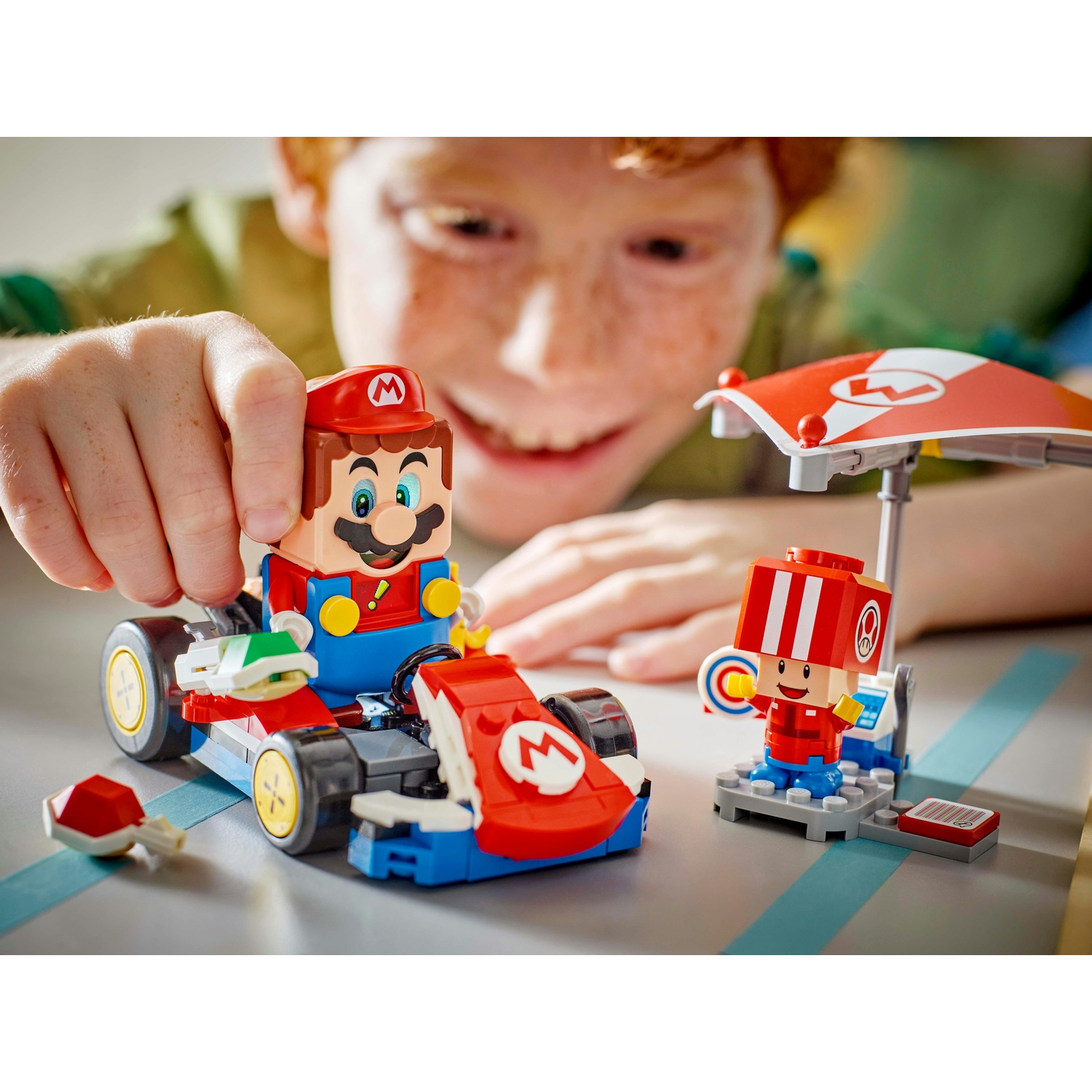 Un bambino gioca con il set Lego Mario Kart. Personaggio di Mario in un kart, un altro personaggio sotto un ombrello rosso. Il bambino sorride.