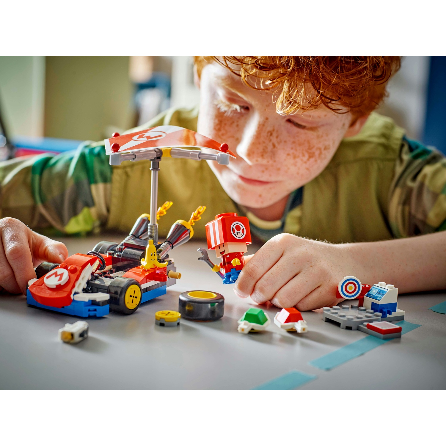 Un ragazzo con le lentiggini gioca con Lego Mario Kart. Ci sono pezzi Lego del kart, personaggi e altri accessori sul tavolo.