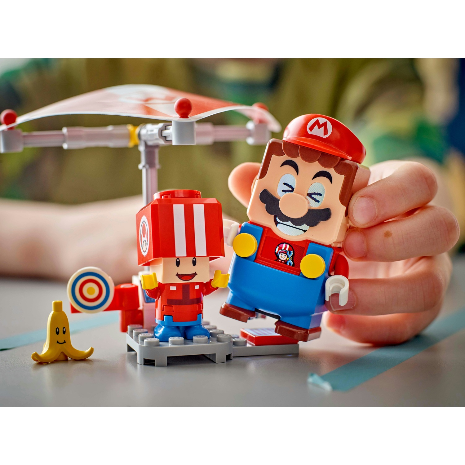 Le figure di Lego Mario e amico sono tenute da una mano. A sinistra una banana gialla e un bersaglio. Un baldacchino sullo sfondo.