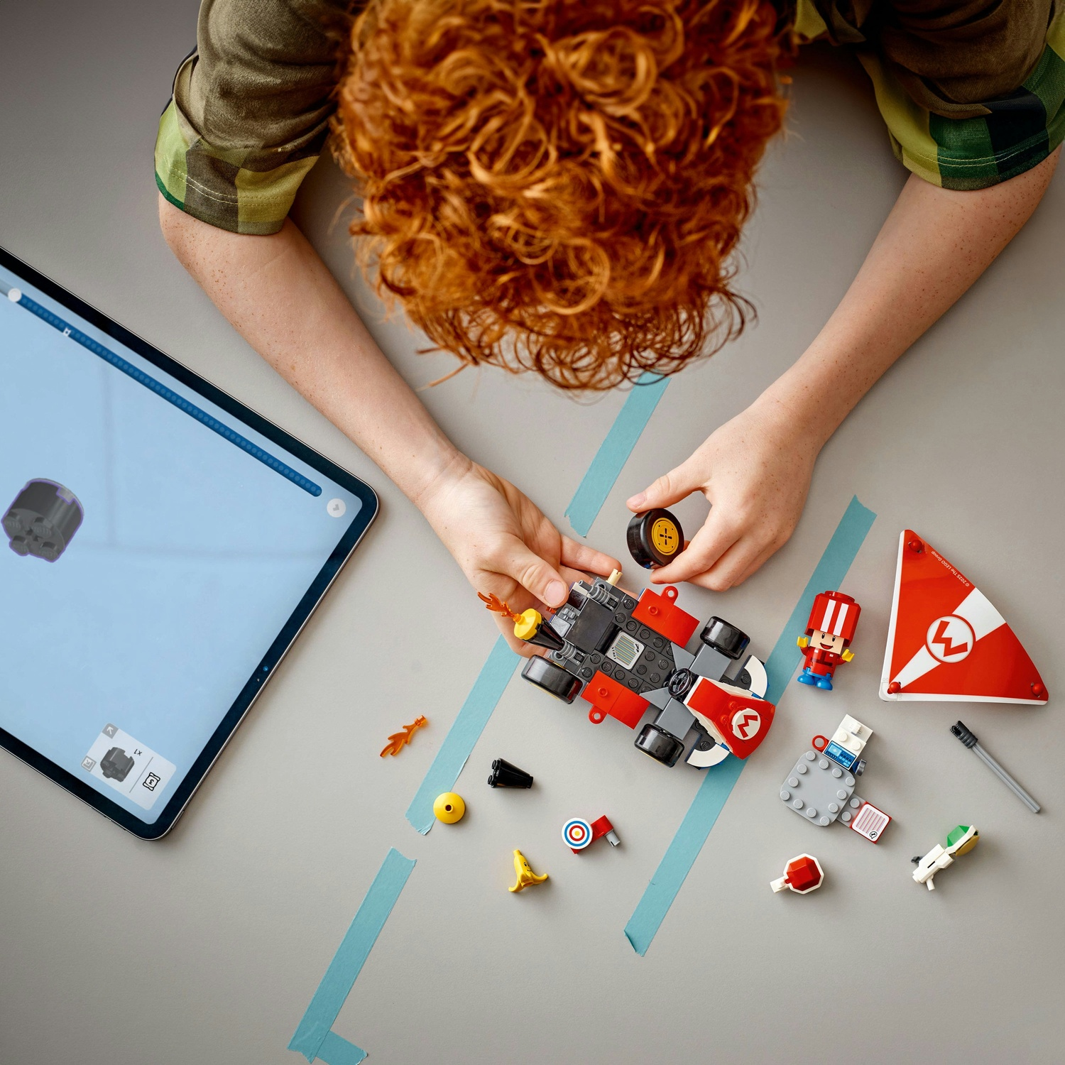 Un bambino dai capelli rossi costruisce un'auto giocattolo con mattoncini Lego. Un tablet mostra le istruzioni. Diversi pezzi Lego sono sparsi.