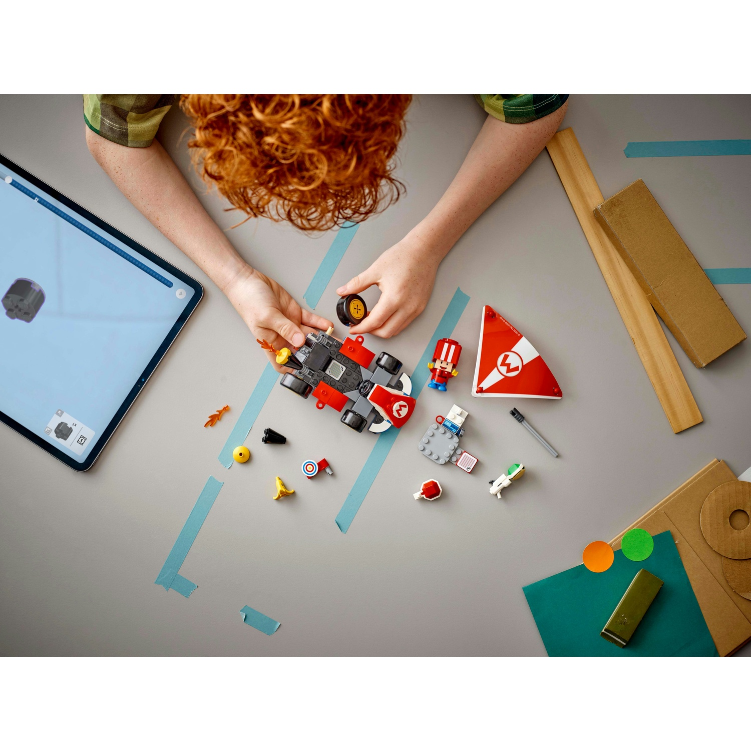 Un bambino sta costruendo un'auto giocattolo con mattoncini LEGO. Vari pezzi LEGO, tra cui una figura, sono sparsi attorno a un tablet.