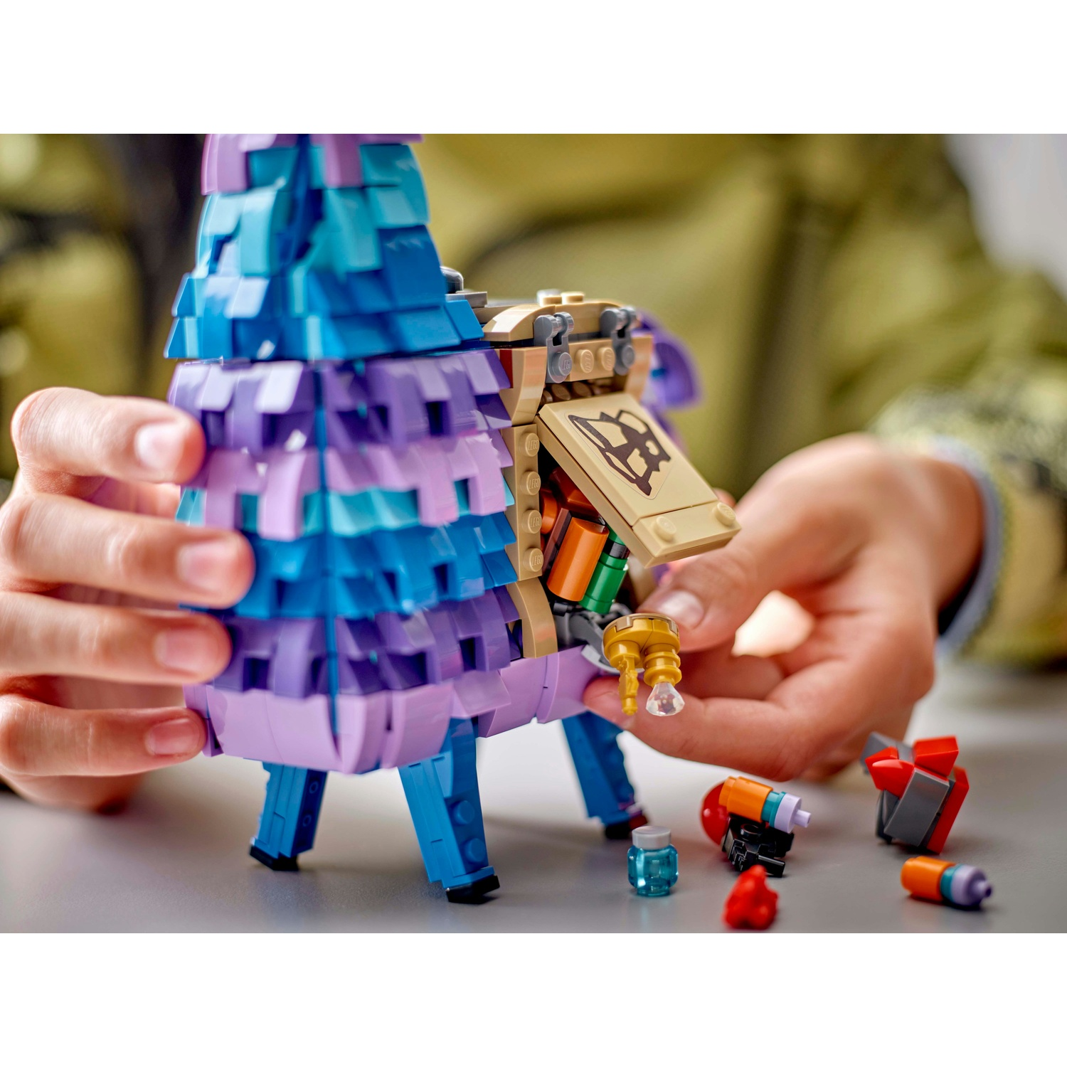 Un lama pignatta Lego con parti blu, viola e beige è tenuto in mano. Piccoli accessori Lego sono sparsi intorno al lama.