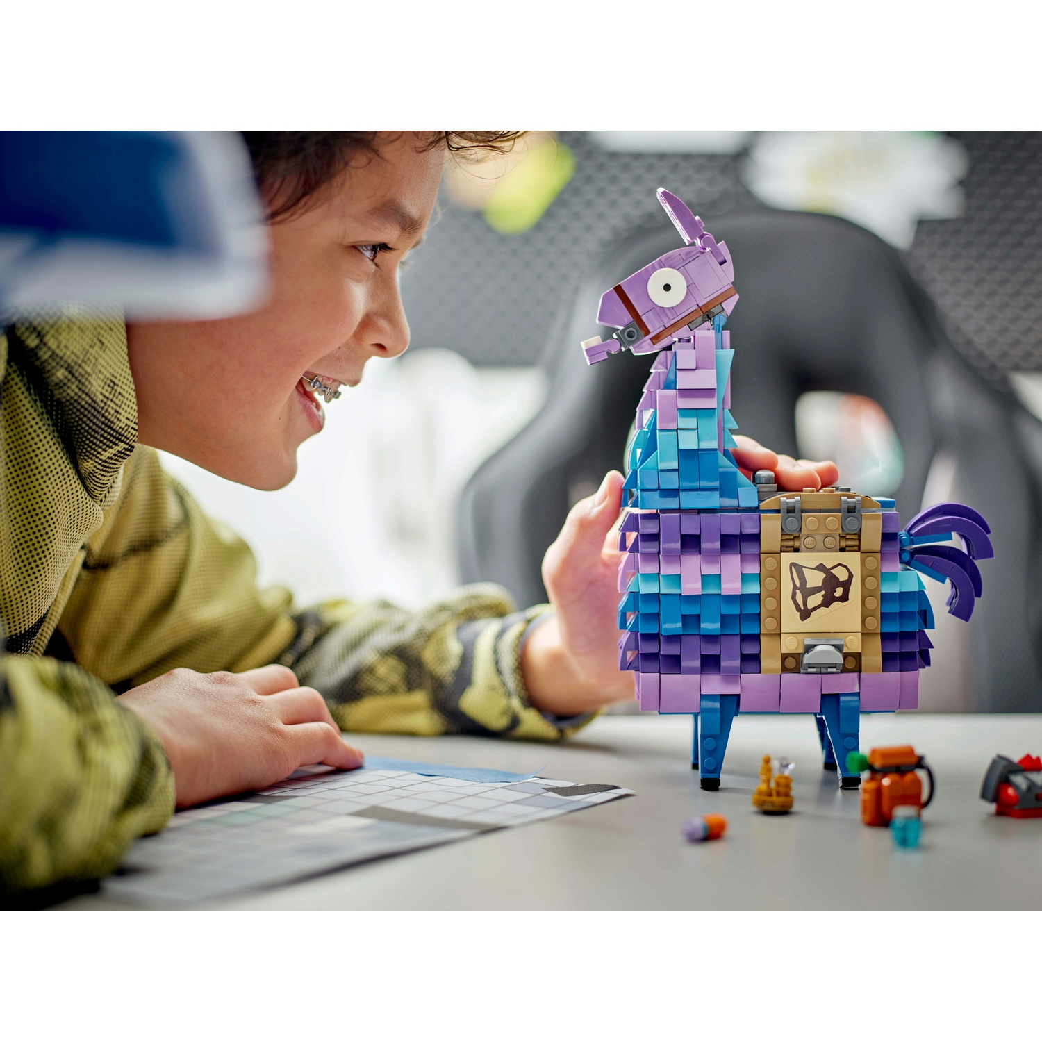 Un ragazzo con l'apparecchio sorride a un lama Lego. Il lama è blu, viola e marrone. Piccoli oggetti Lego sono sul tavolo.