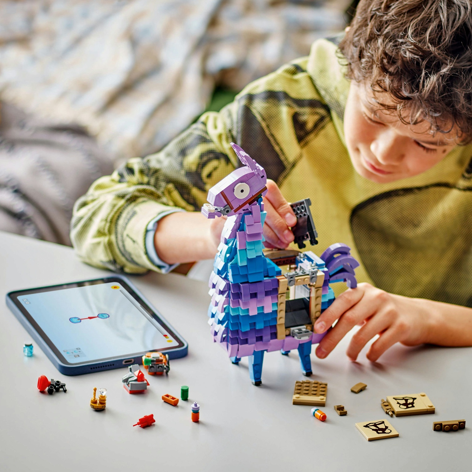 Un ragazzo assembla un lama Lego con un tablet che mostra le istruzioni. Vari pezzi Lego sono sul tavolo bianco.