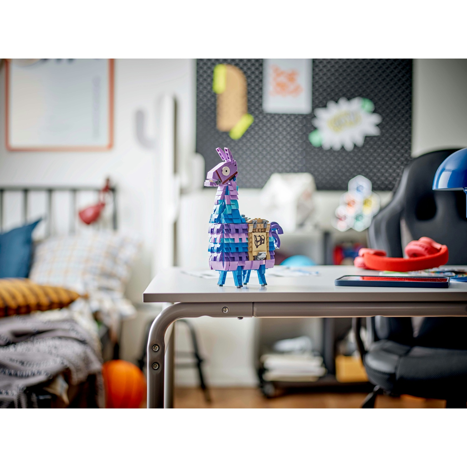 Un lama Lego è su una scrivania. Dietro c'è una lavagna nera, un letto e una sedia. Il lama è viola, blu e bianco.