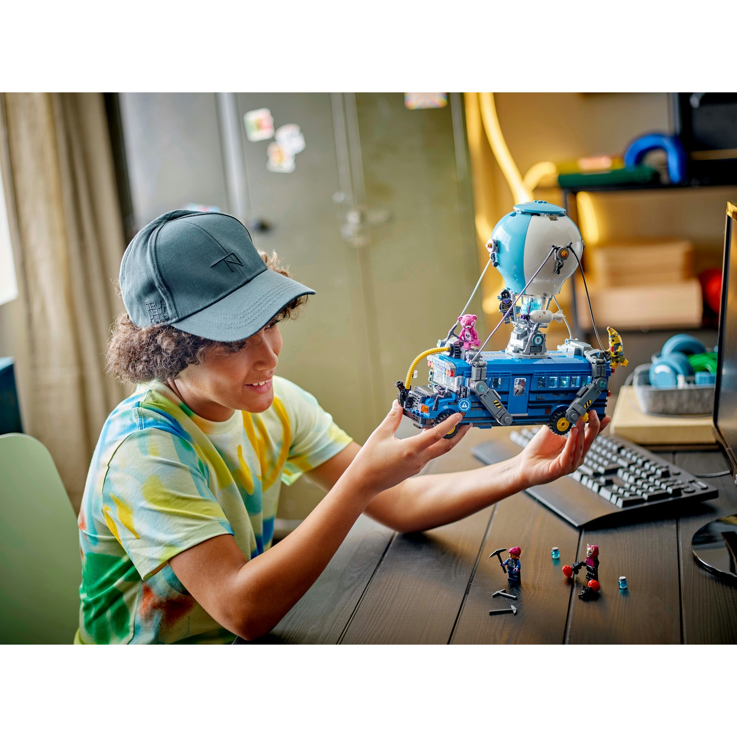 Una persona tiene in mano un autobus LEGO blu con un palloncino bianco e blu sopra. C'è un computer e alcune statuette sul tavolo.