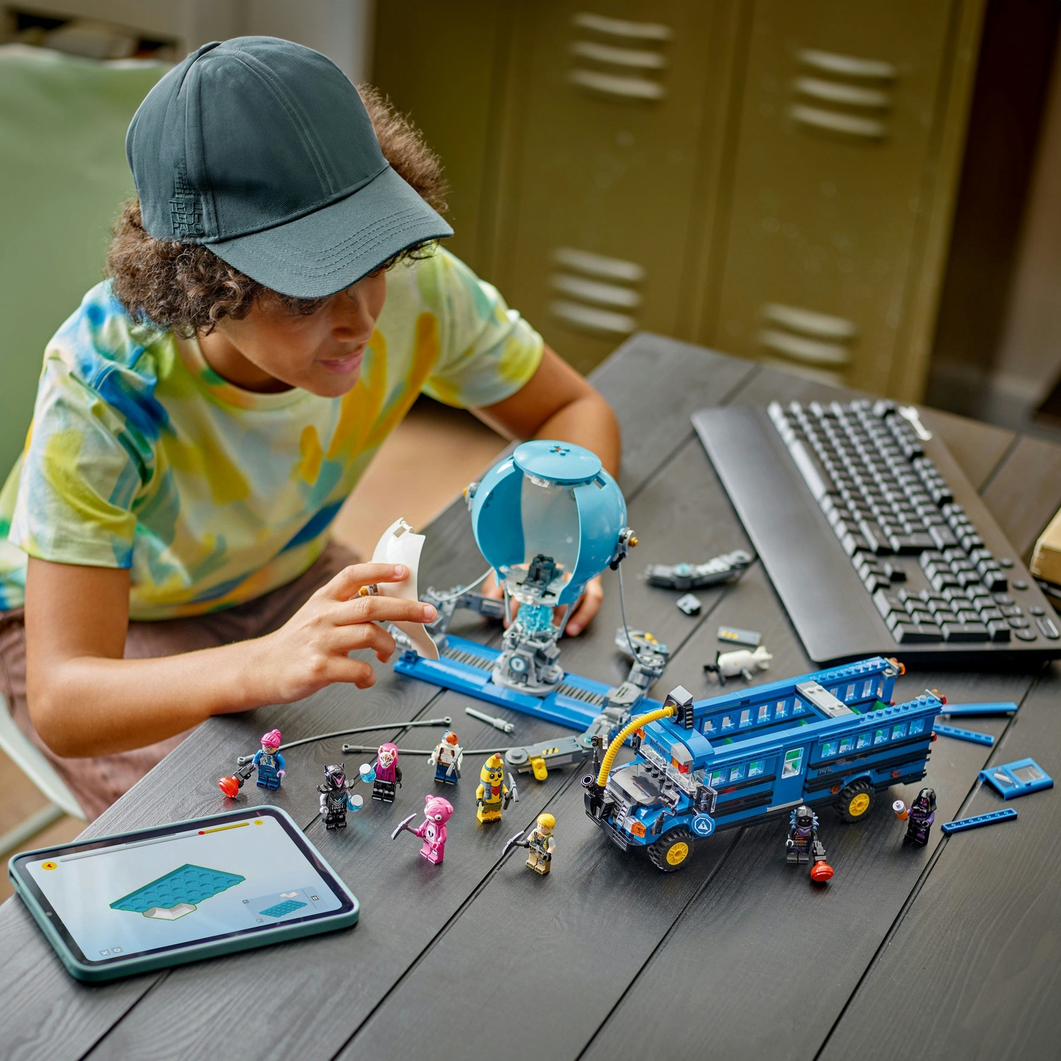 Una persona costruisce un set Lego con un autobus blu e minifigure. Un tablet mostra le istruzioni. Una tastiera è visibile sul tavolo.