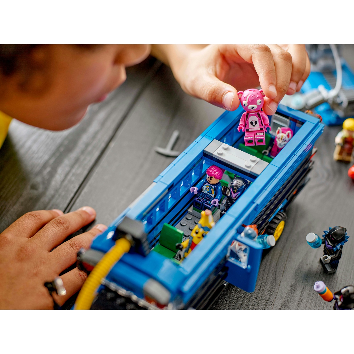 La mano di un bambino posiziona un personaggio Lego a forma di orso rosa in un set Lego di autobus blu, con altri personaggi dentro e intorno.