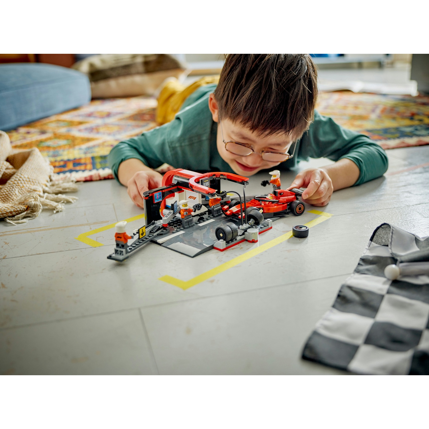 Un ragazzo con gli occhiali gioca con un'auto da corsa Lego rossa e un pit stop sul pavimento. Una bandiera a scacchi  è vicina.