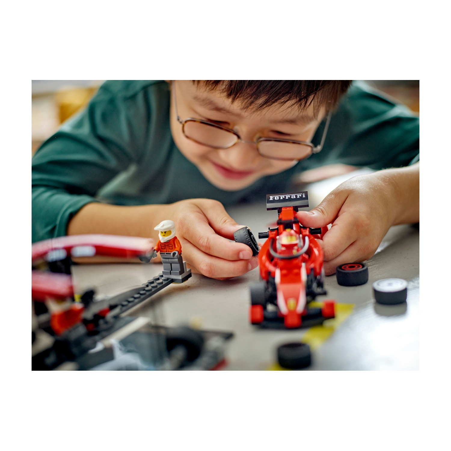 Un bambino con gli occhiali gioca con un'auto da corsa Ferrari Lego rossa. Statuine e pneumatici Lego su un tavolo.