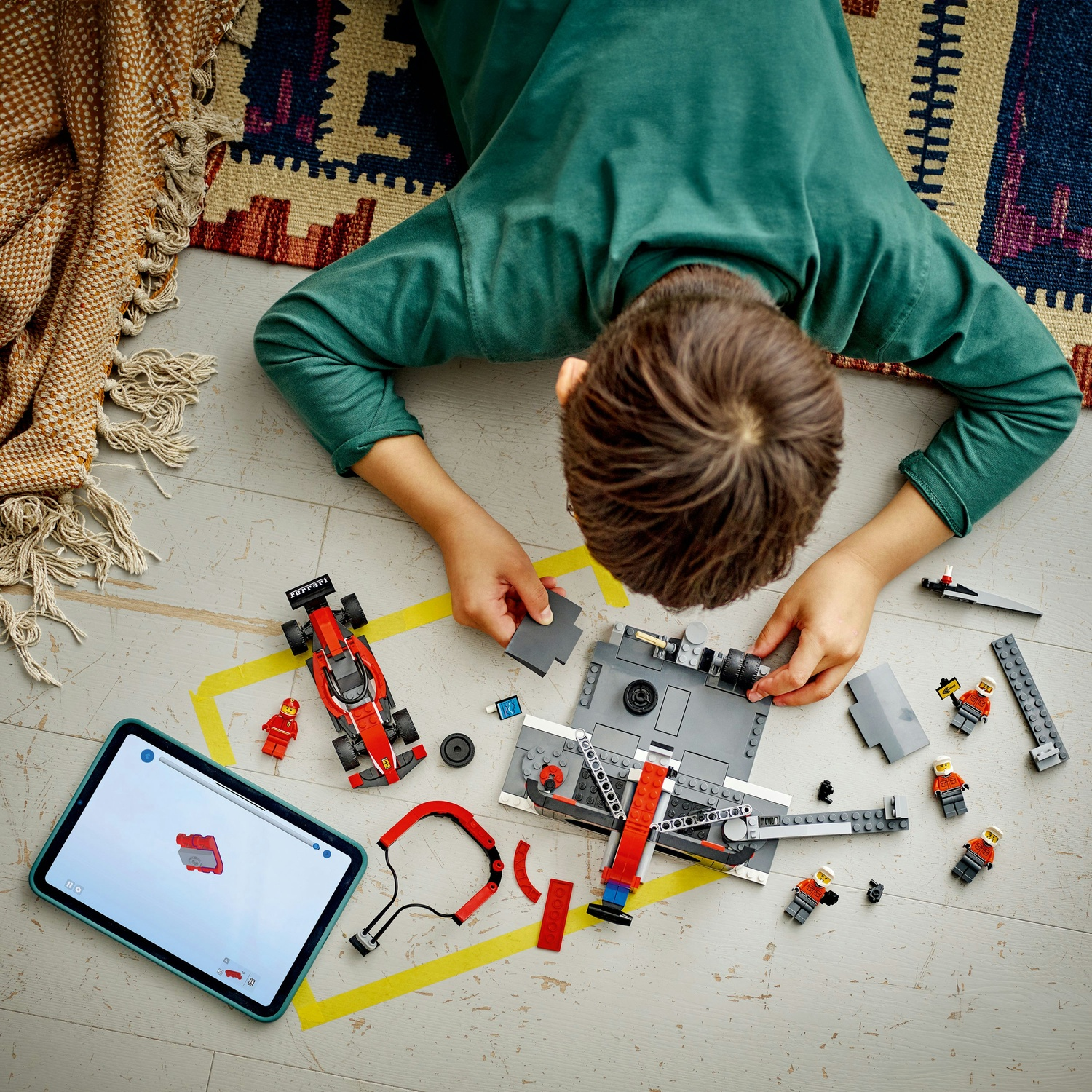 Un bambino sta costruendo un set Lego con un tablet che mostra le istruzioni. Un'auto da corsa rossa e diverse figure Lego sono visibili.