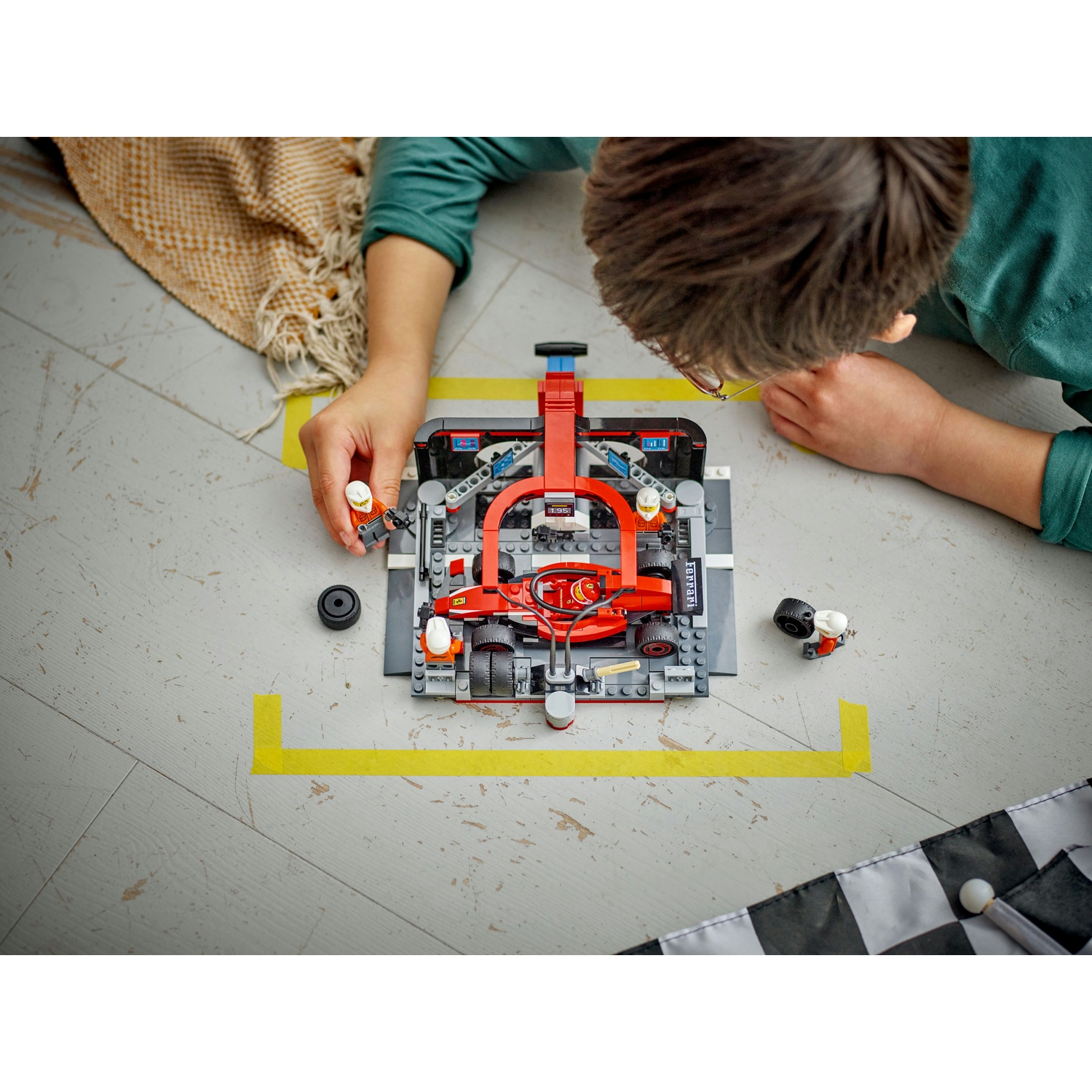 Un bambino assembla un'auto da corsa Lego rossa, con un team di box, su un pavimento grigio con segni di nastro giallo. Una coperta e una bandiera a scacchi sono vicine.