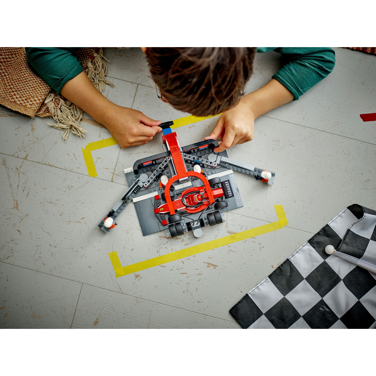 Una persona sta costruendo un'auto da corsa Lego rossa e grigia su una superficie grigia con nastro giallo. Una bandiera a scacchi è nell'angolo.