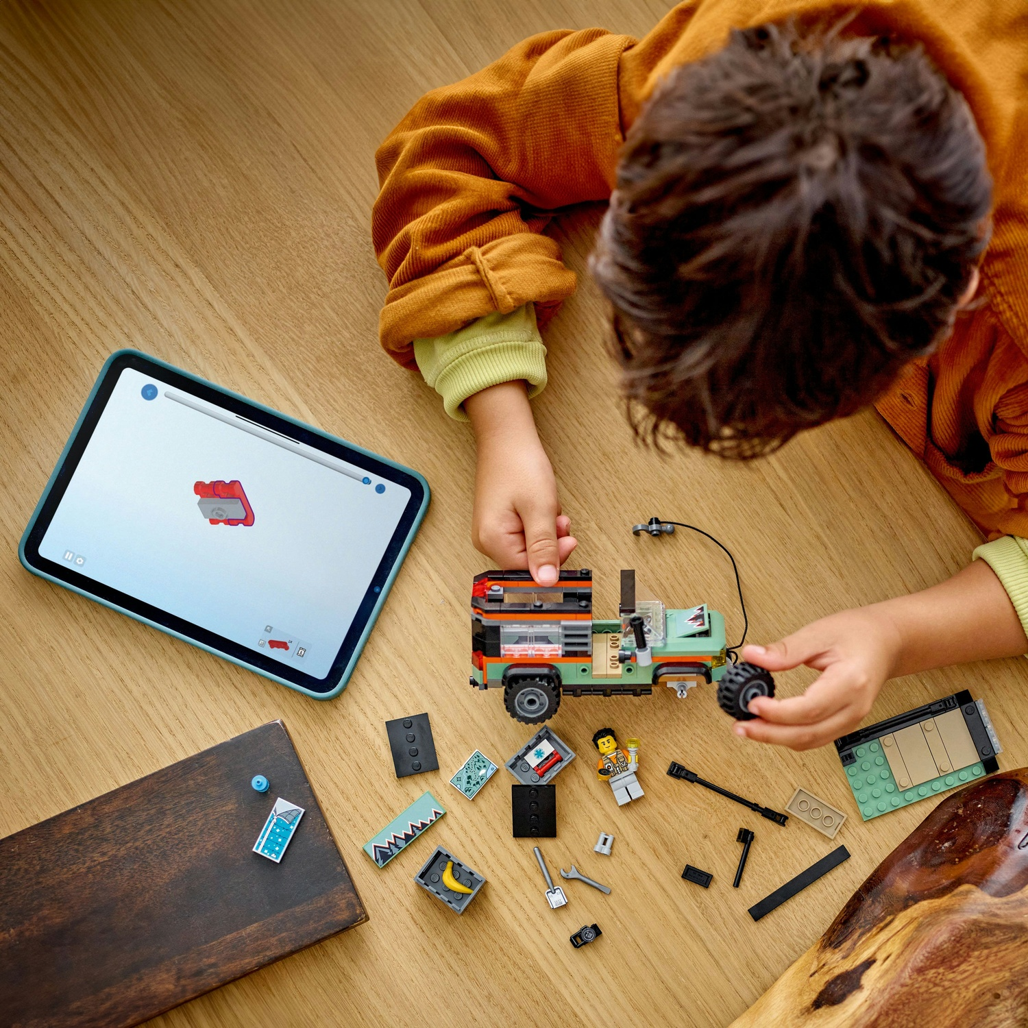 Un bambino assembla un'auto Lego su un tavolo di legno, guidato da un tablet. Parti e una minifigure sono sparse intorno all'auto.
