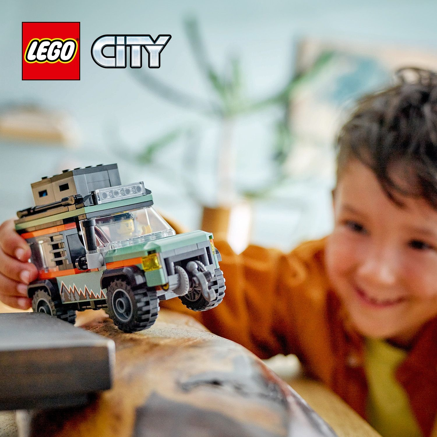 Un bambino sorride, tenendo in mano un veicolo fuoristrada Lego City. Il veicolo è verde, arancione e nero. Un logo Lego è in alto a sinistra.