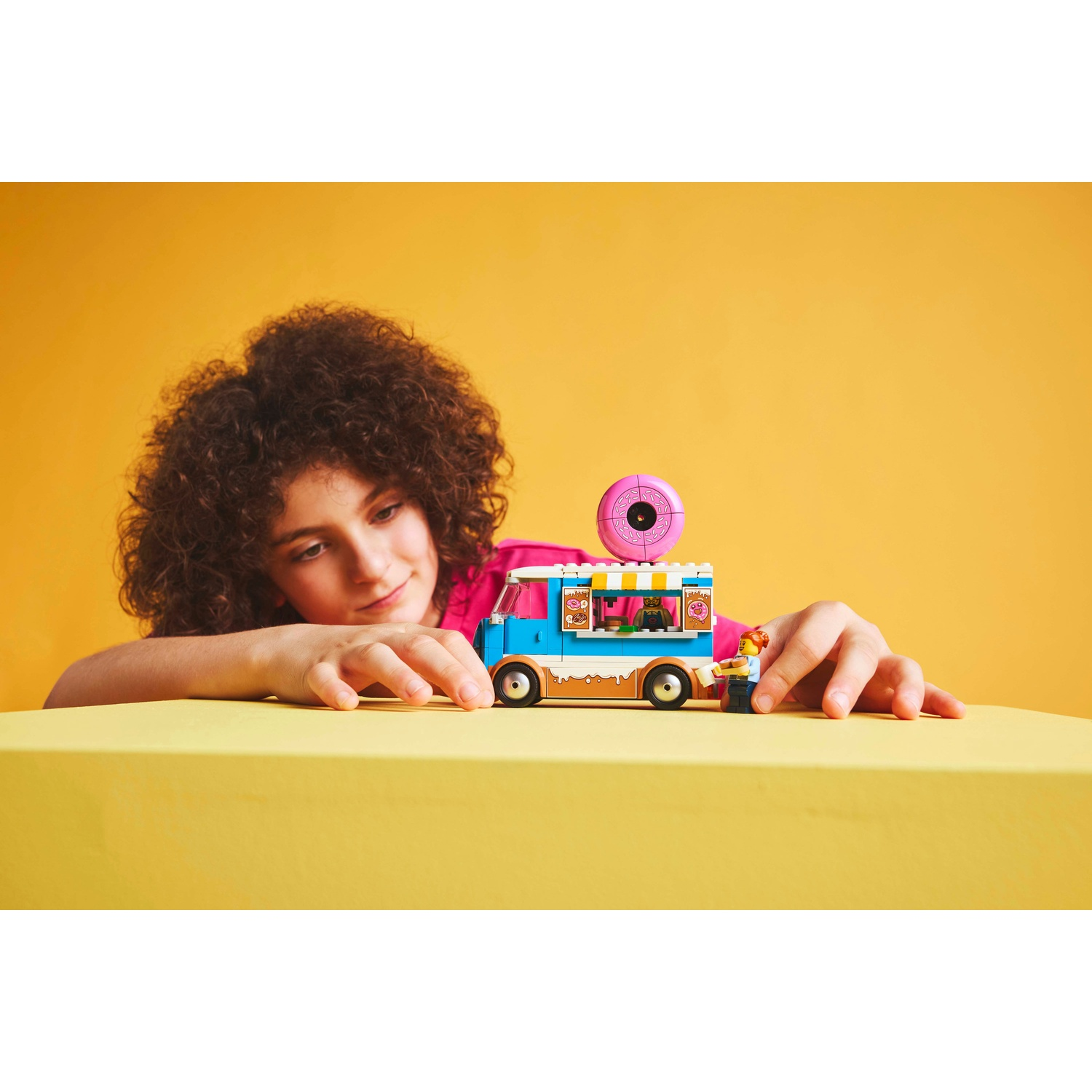 Un bambino guarda un camioncino LEGO. Il camion è blu e marrone. C'è una ciambella rosa sopra. Una minifigure è accanto.