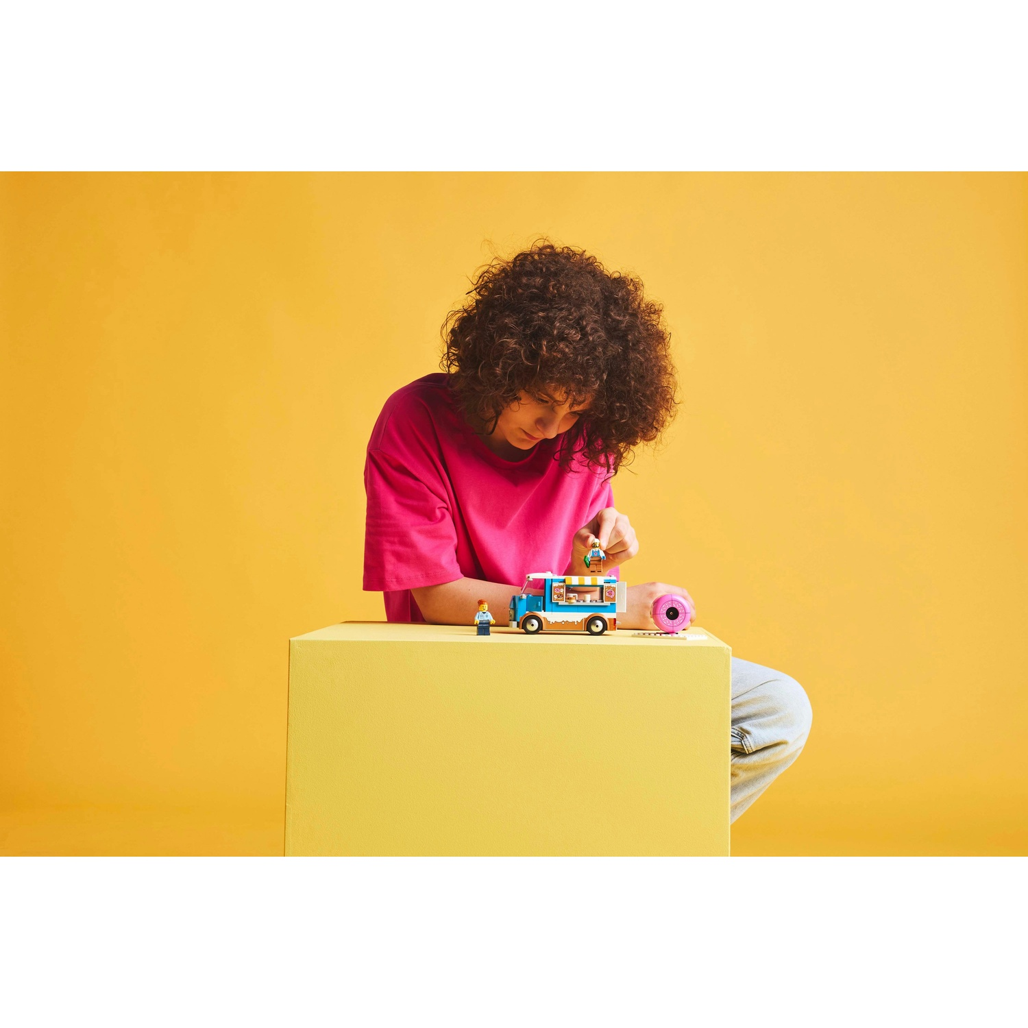 Una persona con una maglietta rosa sta costruendo un camioncino dei gelati Lego su un blocco giallo. Lo sfondo è giallo.