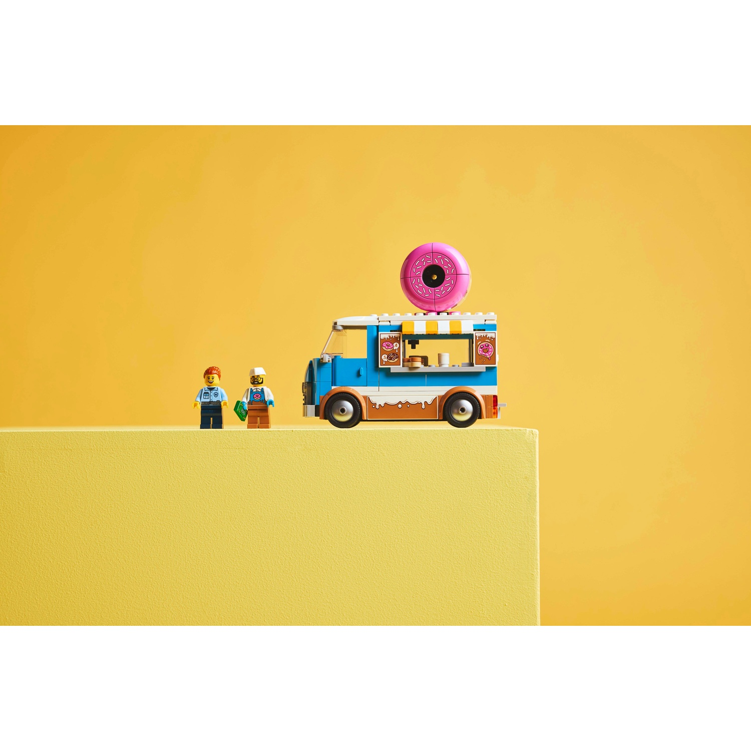 Un camioncino di ciambelle Lego con due minifigure. Una grande ciambella rosa sopra il camion. Lo sfondo è giallo.