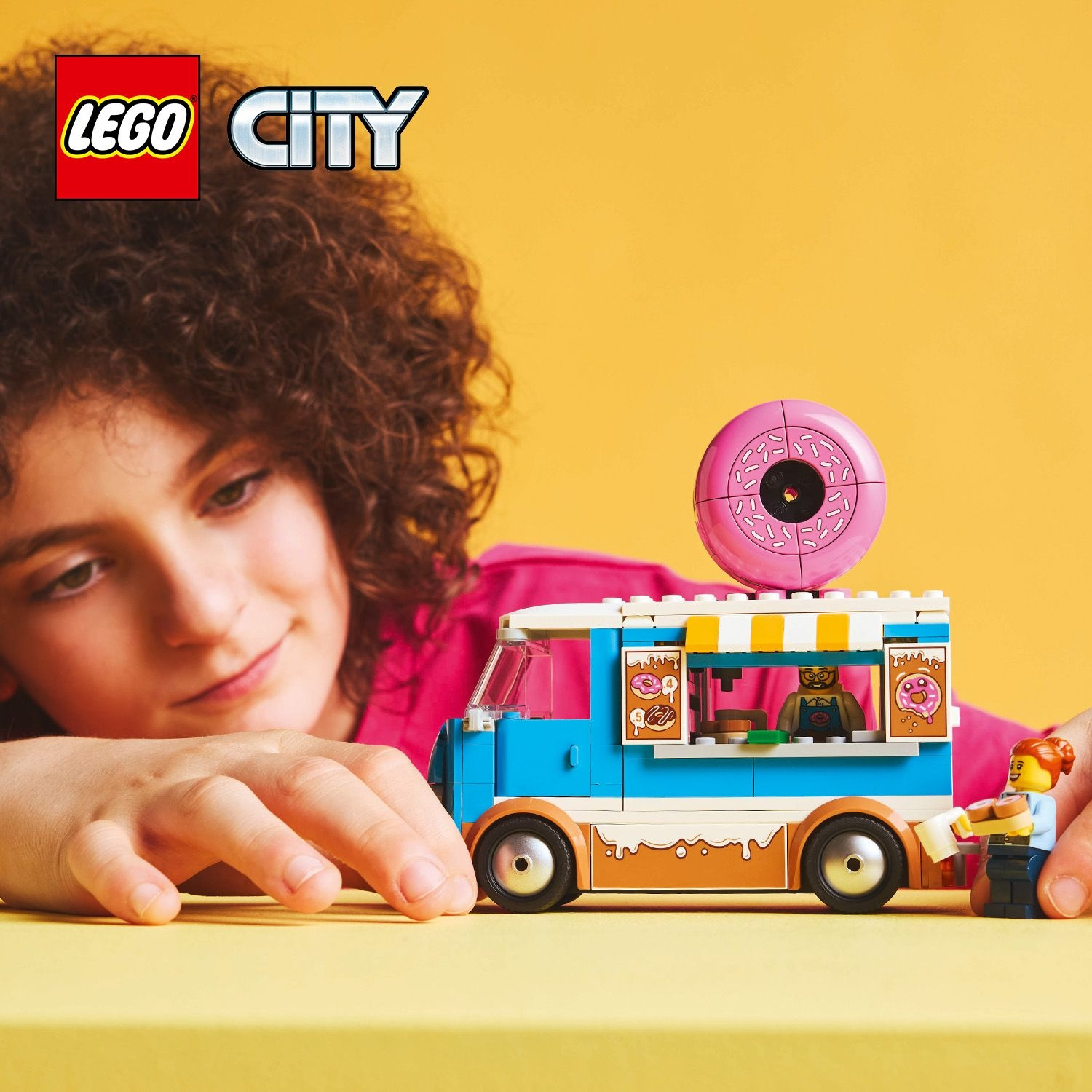 Un bambino guarda un camion Lego City del negozio di ciambelle. Una grande ciambella rosa è in cima. Due personaggi Lego interagiscono con il camion.