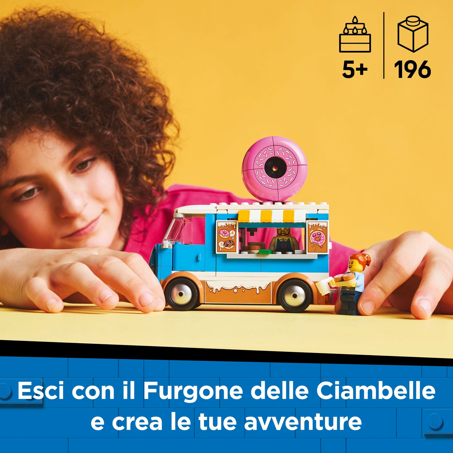 Una ragazza guarda un camioncino di ciambelle Lego con una ciambella rosa sopra. Il camion ha un design blu e marrone. Due personaggi Lego sono vicino al camion.