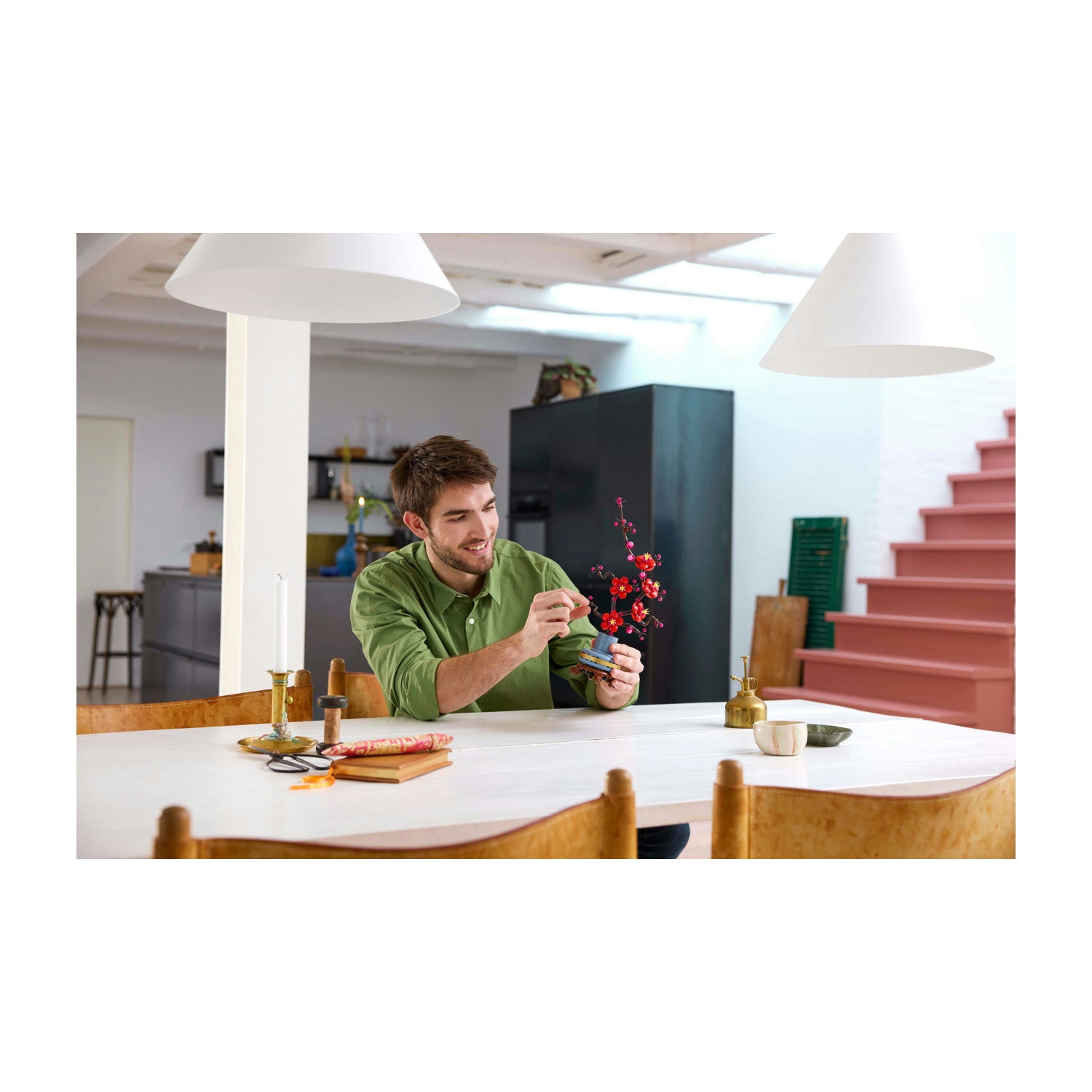 Un uomo con una camicia verde sistema fiori rossi in un vaso su un tavolo bianco. Sono presenti candele e ciotole. Scala sullo sfondo.