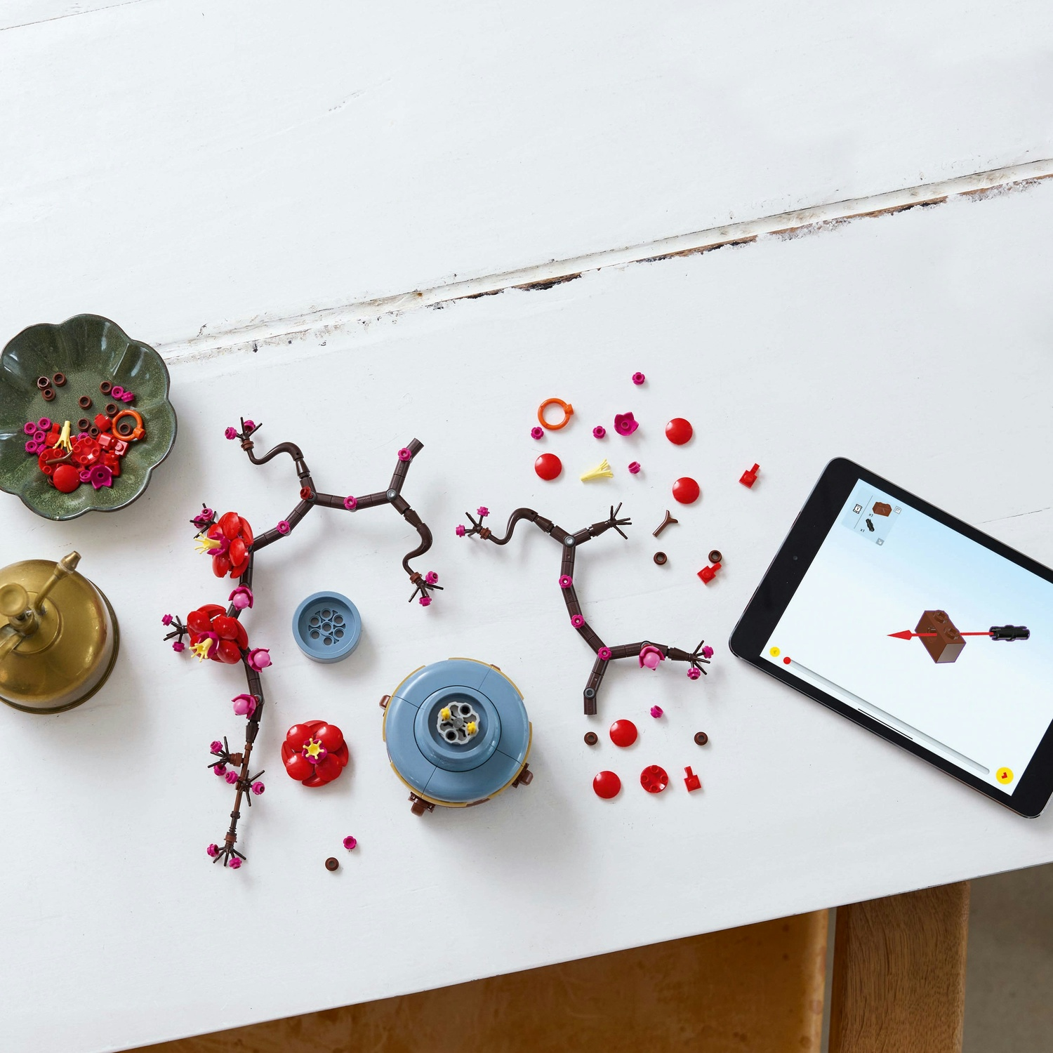 Componenti del set Lego su tavolo bianco. Include fiori rossi, rami marroni, base blu e tablet con istruzioni.