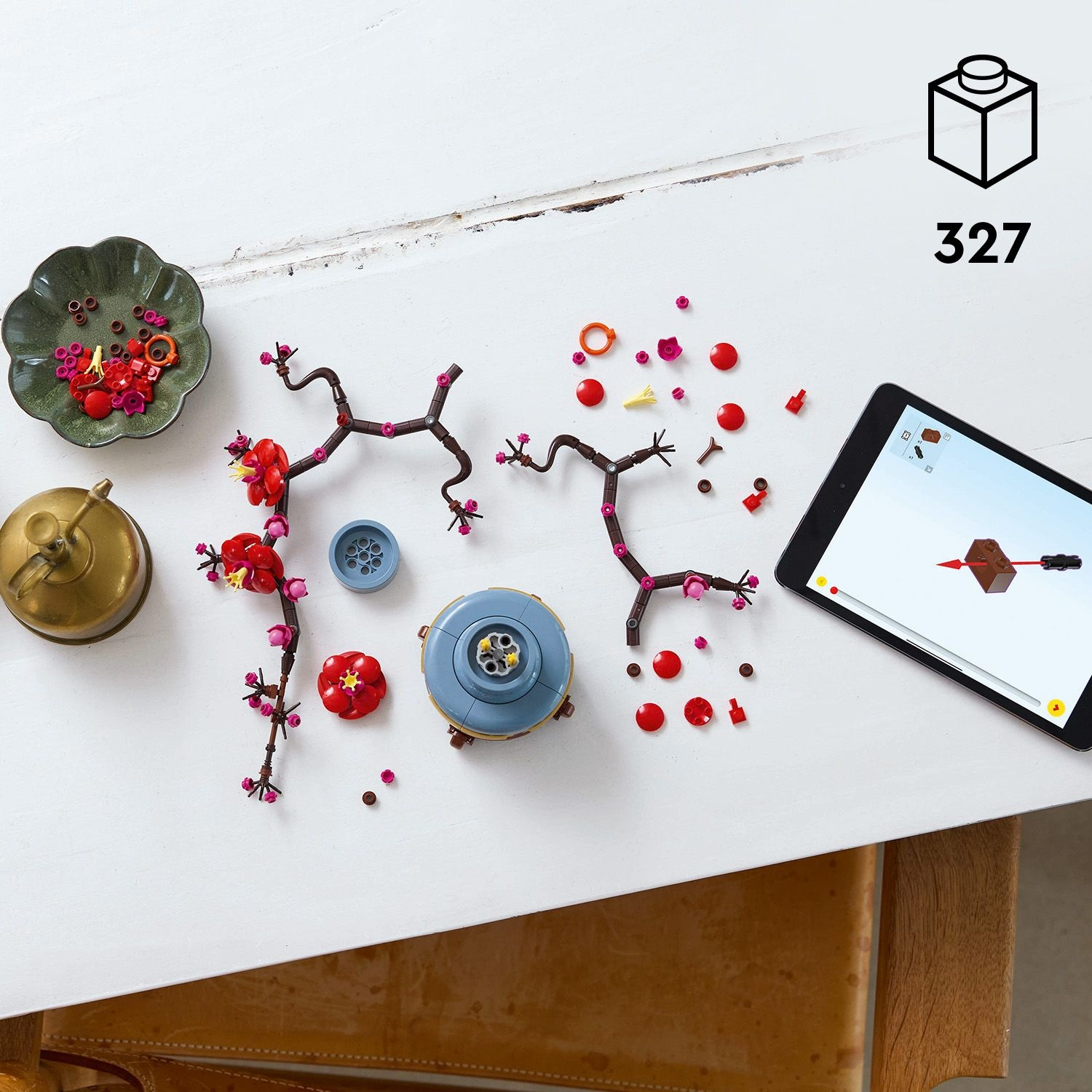 Un tavolo bianco con un set Lego con fiori e istruzioni su un tablet. Altri oggetti includono un contenitore di ottone e una ciotola verde.