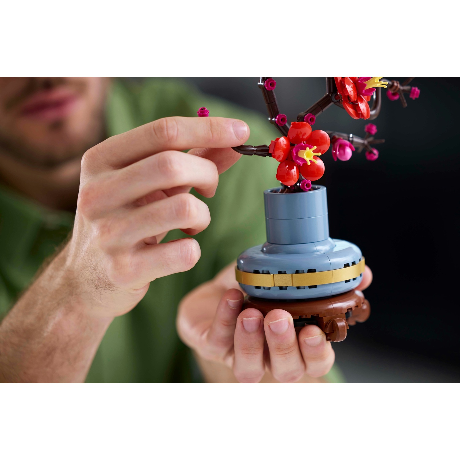 Una persona tiene in mano una composizione floreale Lego. Ha fiori rossi, steli marroni e un vaso azzurro. Lo sfondo è scuro.