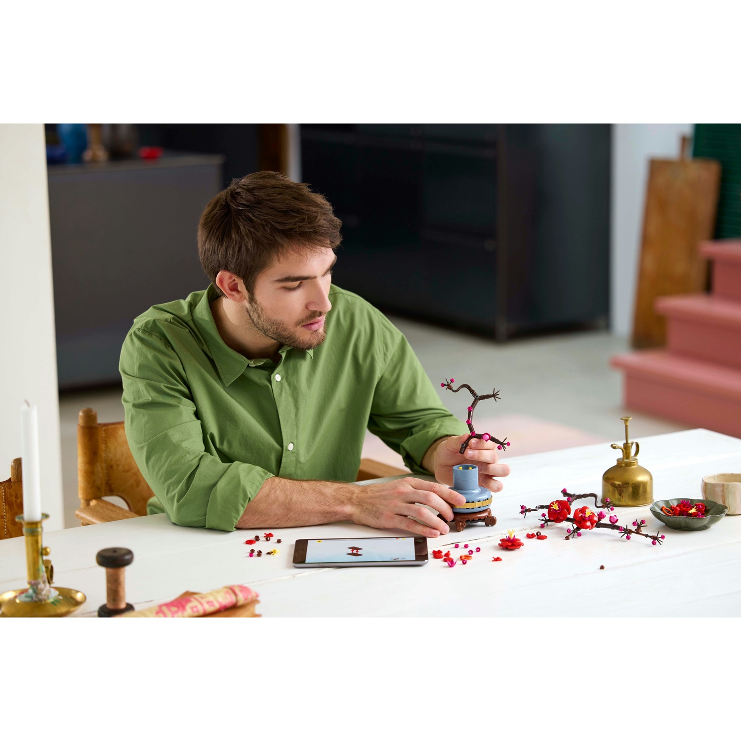 Un uomo con una camicia verde sta assemblando un bonsai Lego su un tavolo bianco. Sono presenti vari pezzi e strumenti Lego.
