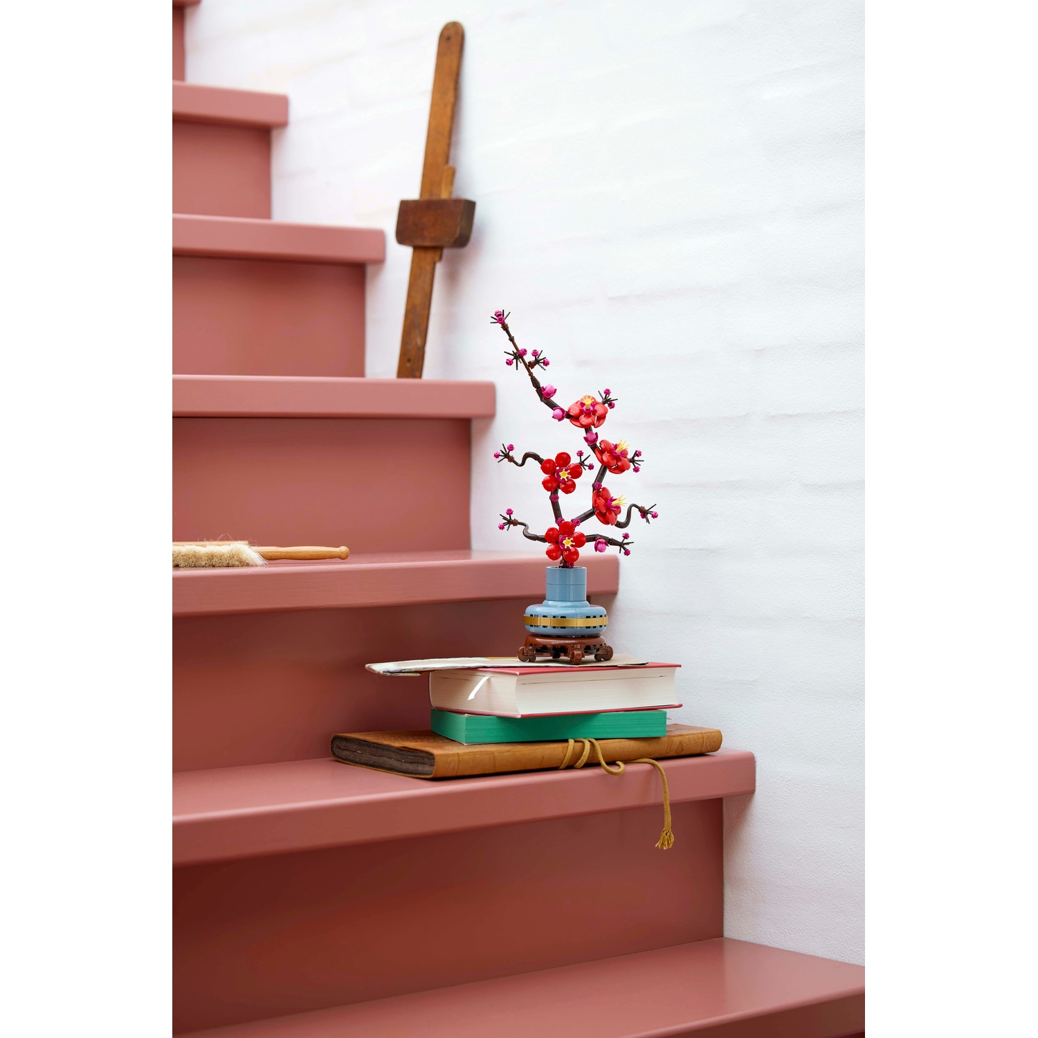 Scale rosa con libri e un vaso di fiori rossi. Uno strumento di legno poggia sulle scale. Muro bianco strutturato sullo sfondo.
