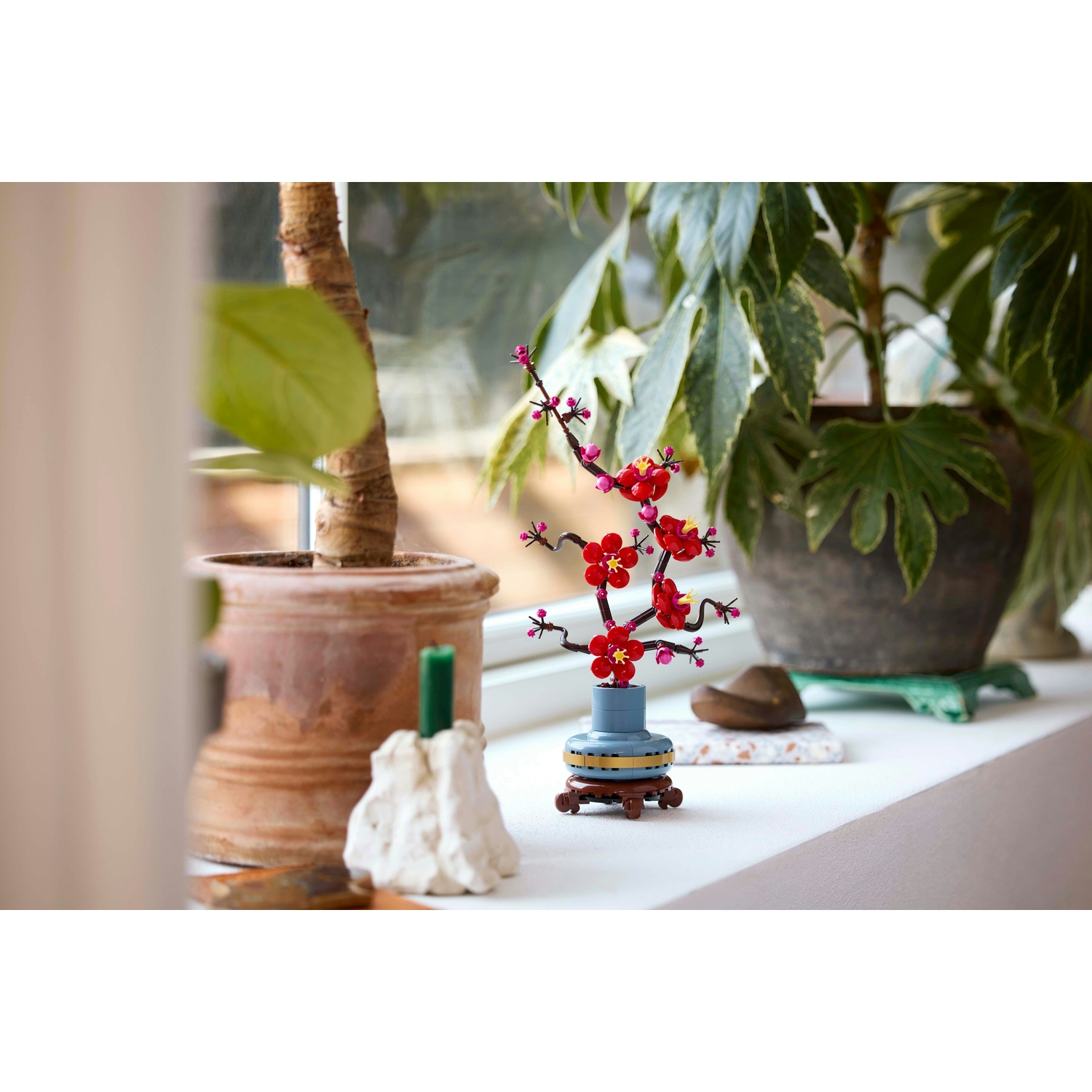 Un bonsai Lego con fiori rossi e un vaso blu è appoggiato su un davanzale accanto a piante in vaso e un portacandele bianco.