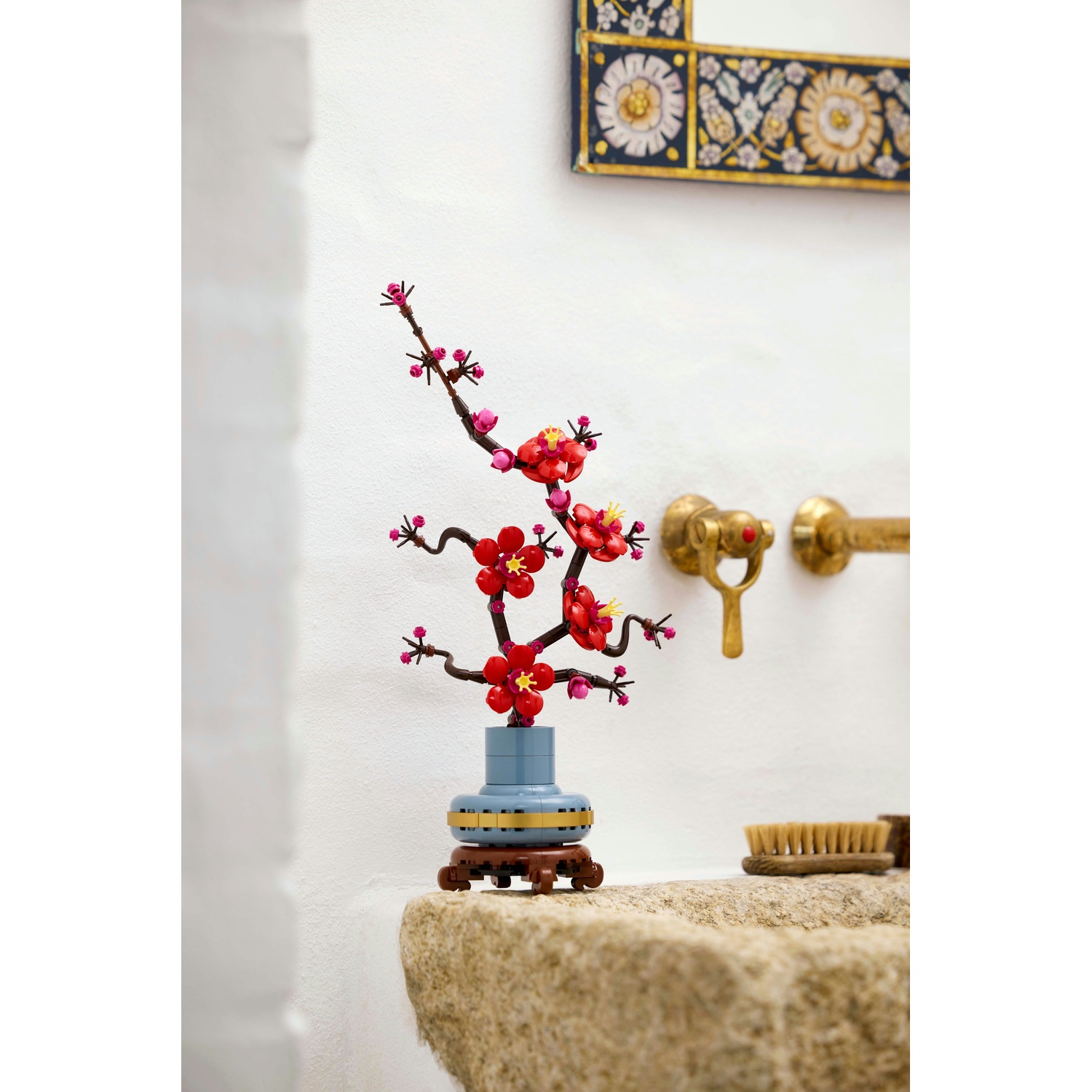 Composizione floreale decorativa con fiori rossi e rami marroni, in un vaso blu, su un lavabo in pietra, vicino a rubinetti dorati e uno specchio.