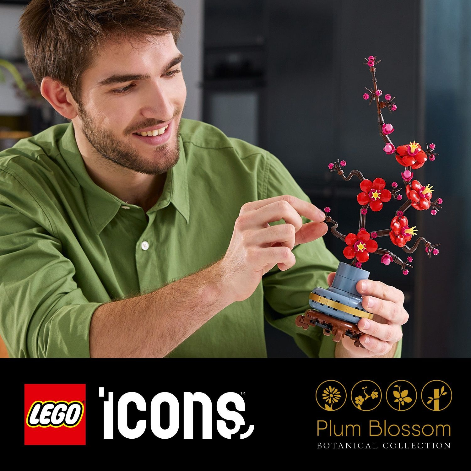 Un uomo tiene in mano un fiore di prugno LEGO. Il fiore ha fiori rossi e steli marroni. L'uomo indossa una camicia verde e sorride.