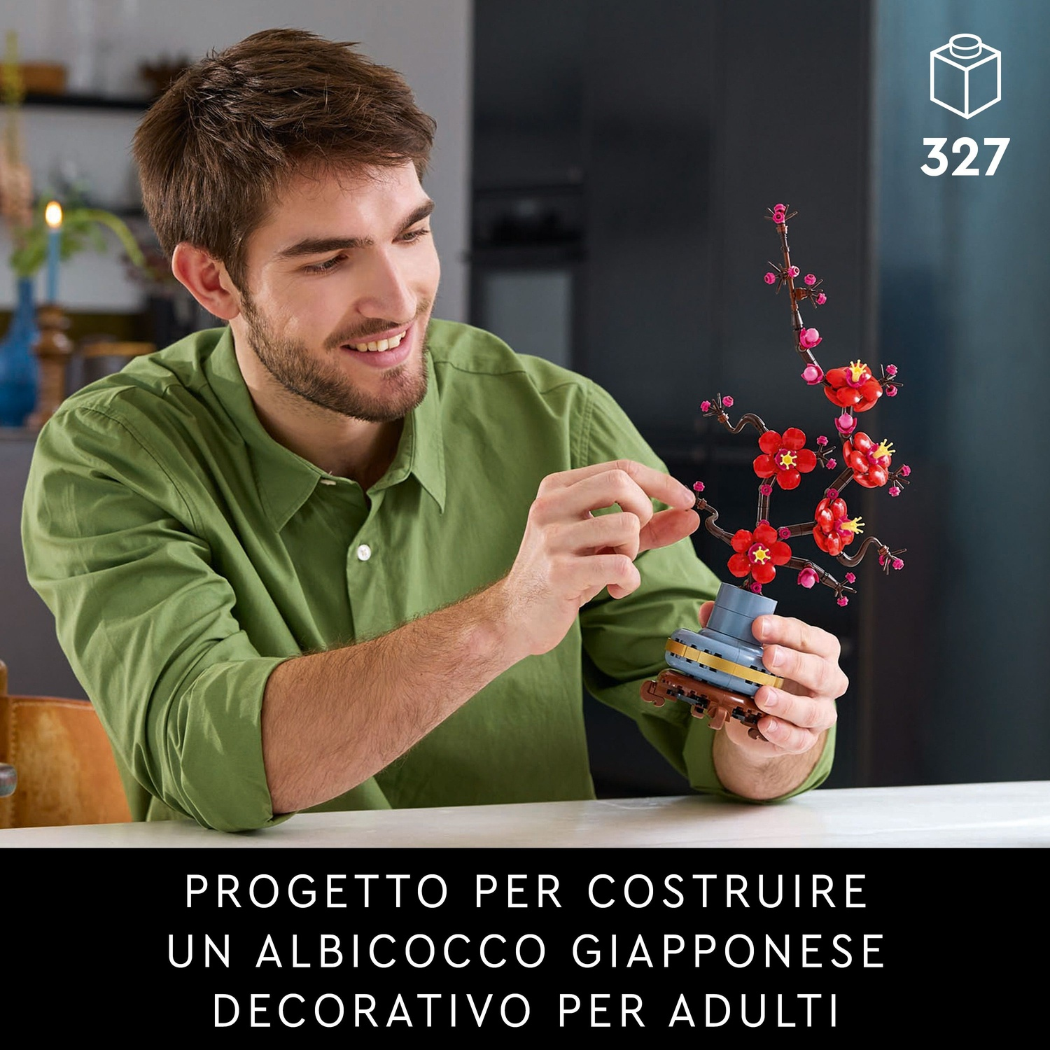 Un uomo tiene in mano un set Lego di fiori di ciliegio. Il set ha fiori rossi e rami marroni. L'uomo indossa una camicia verde e sorride.
