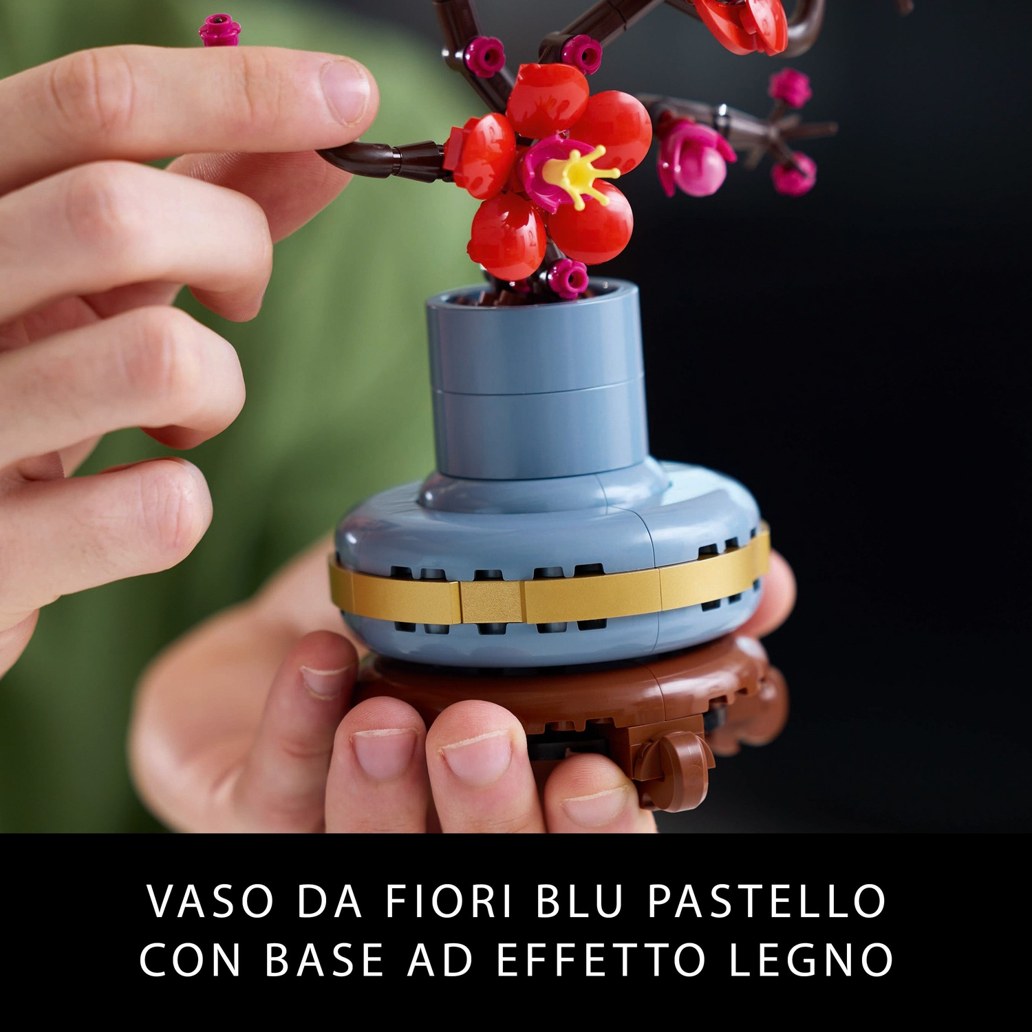 Mani che tengono un vaso blu pastello con base in legno, contenente una composizione floreale con fiori rossi e rosa.