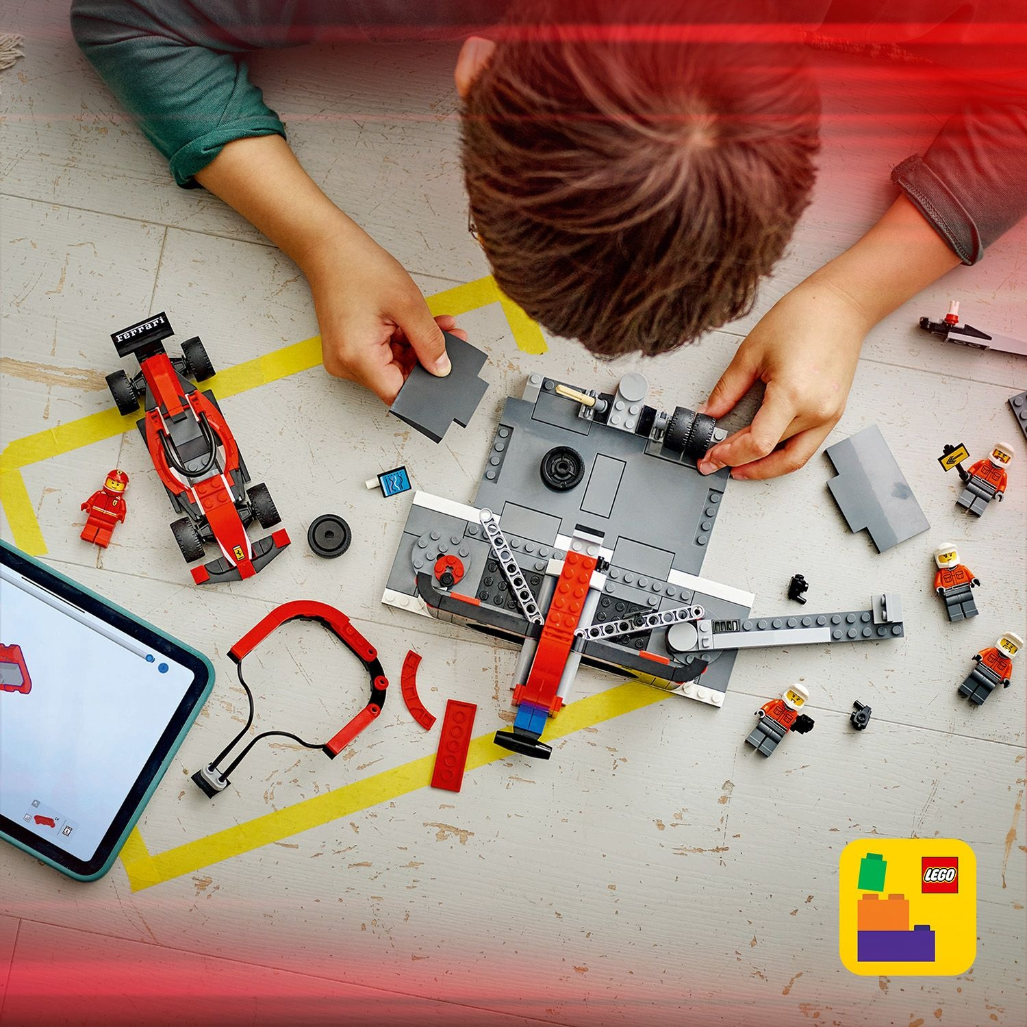 Un bambino assembla un'auto da corsa Lego sul pavimento. Pezzi, tablet con istruzioni e personaggi Lego sono sparsi.