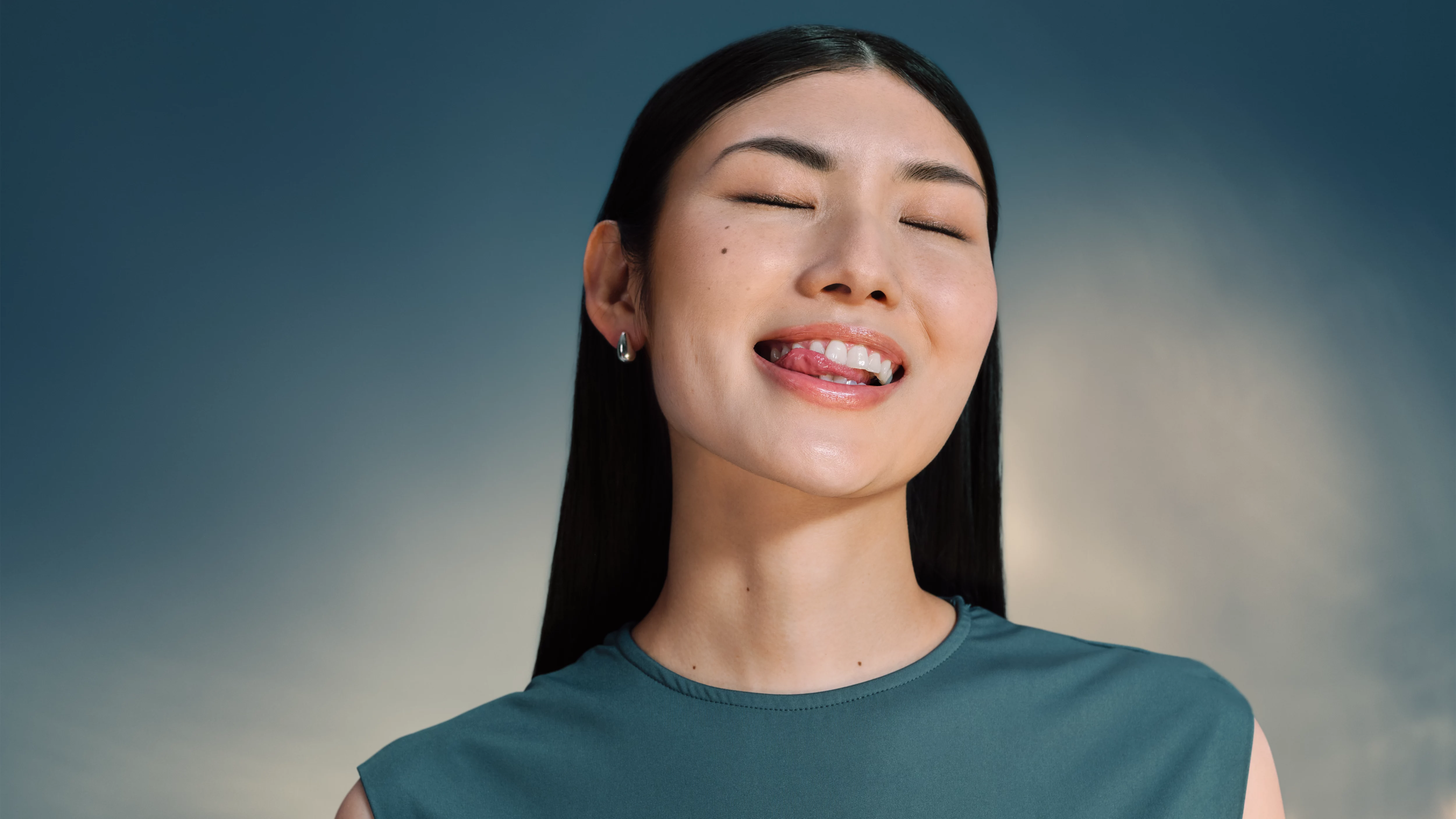 Kobieta z zamkniętymi oczami wystawia język. Ma na sobie koszulę w kolorze morskim i srebrny kolczyk. Na tle niebo.