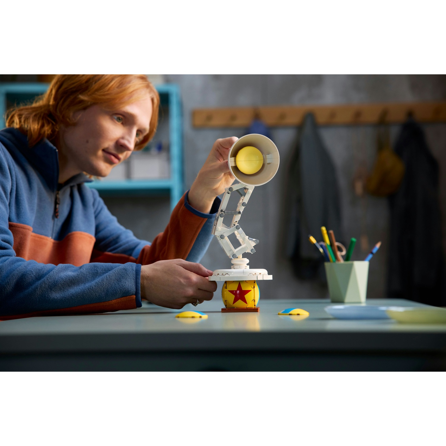 Un uomo dai capelli rossi assembla un modello di lampada Pixar. La lampada è gialla e bianca. Lo sfondo mostra una scrivania e una mensola.