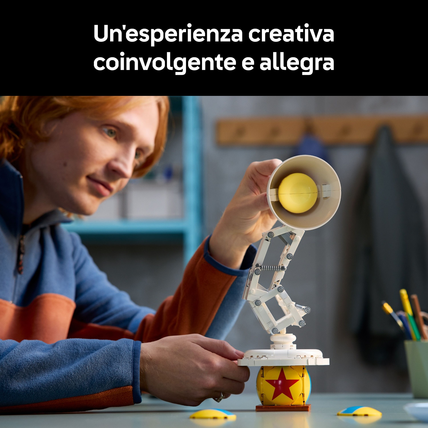 Una persona tiene in mano un modello di lampada da scrivania bianca. La lampada ha un paralume giallo e poggia su una palla gialla. La persona indossa una camicia blu e arancione.
