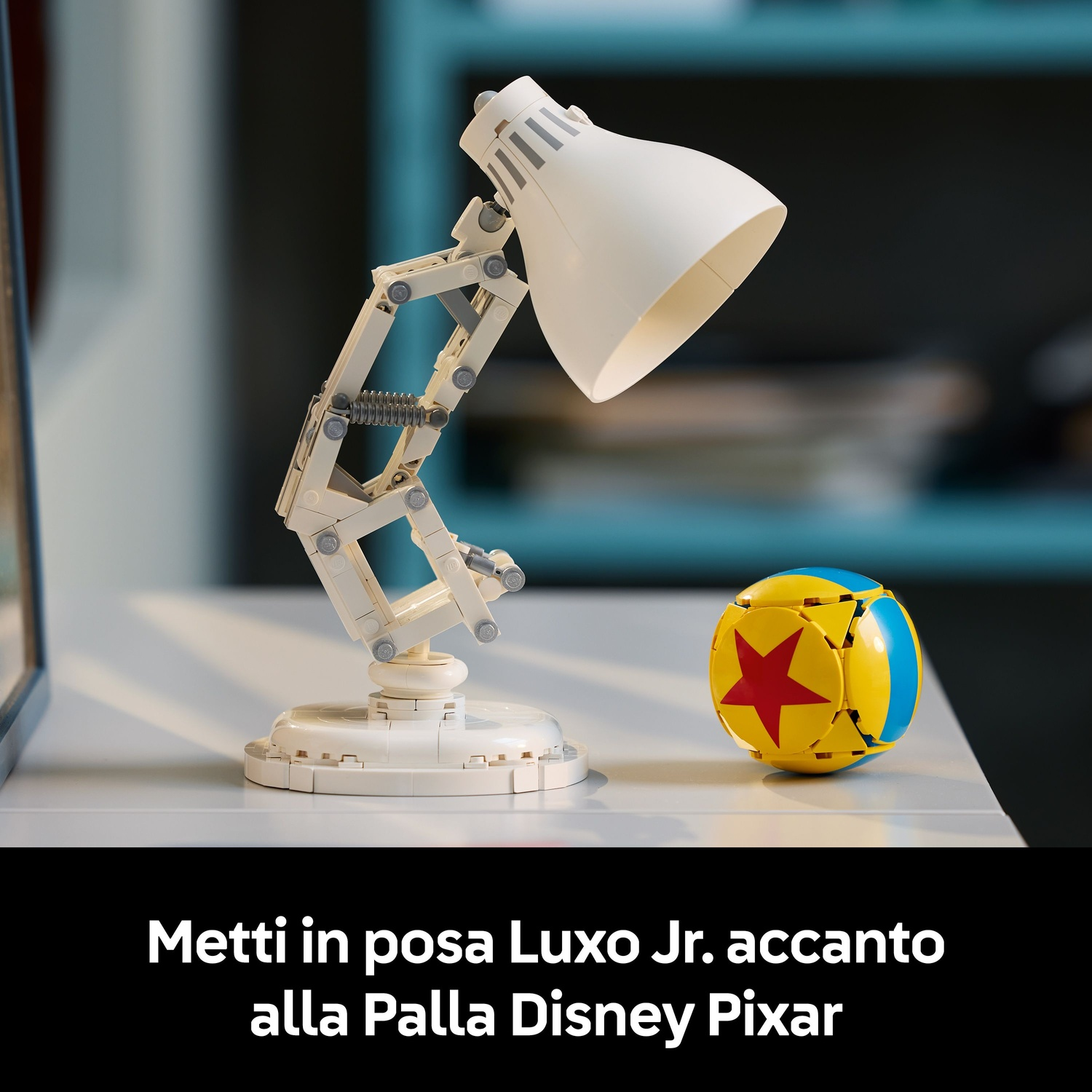 Una lampada da scrivania Lego bianca e una palla Pixar sono su una superficie bianca. Lo sfondo è sfocato, con tonalità scure e verde acqua.