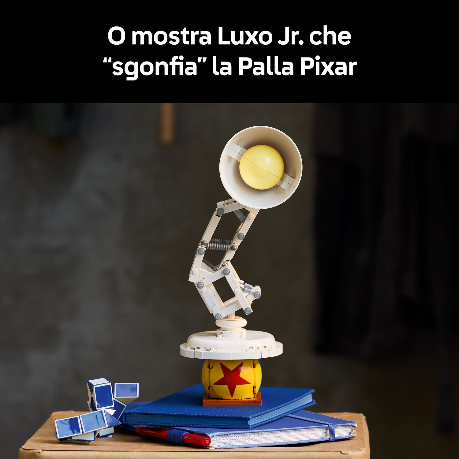 Una lampada Lego Luxo Jr. è su una scrivania con una palla, libri e una penna. La lampada è bianca, ha una luce gialla ed è diretta verso la palla.