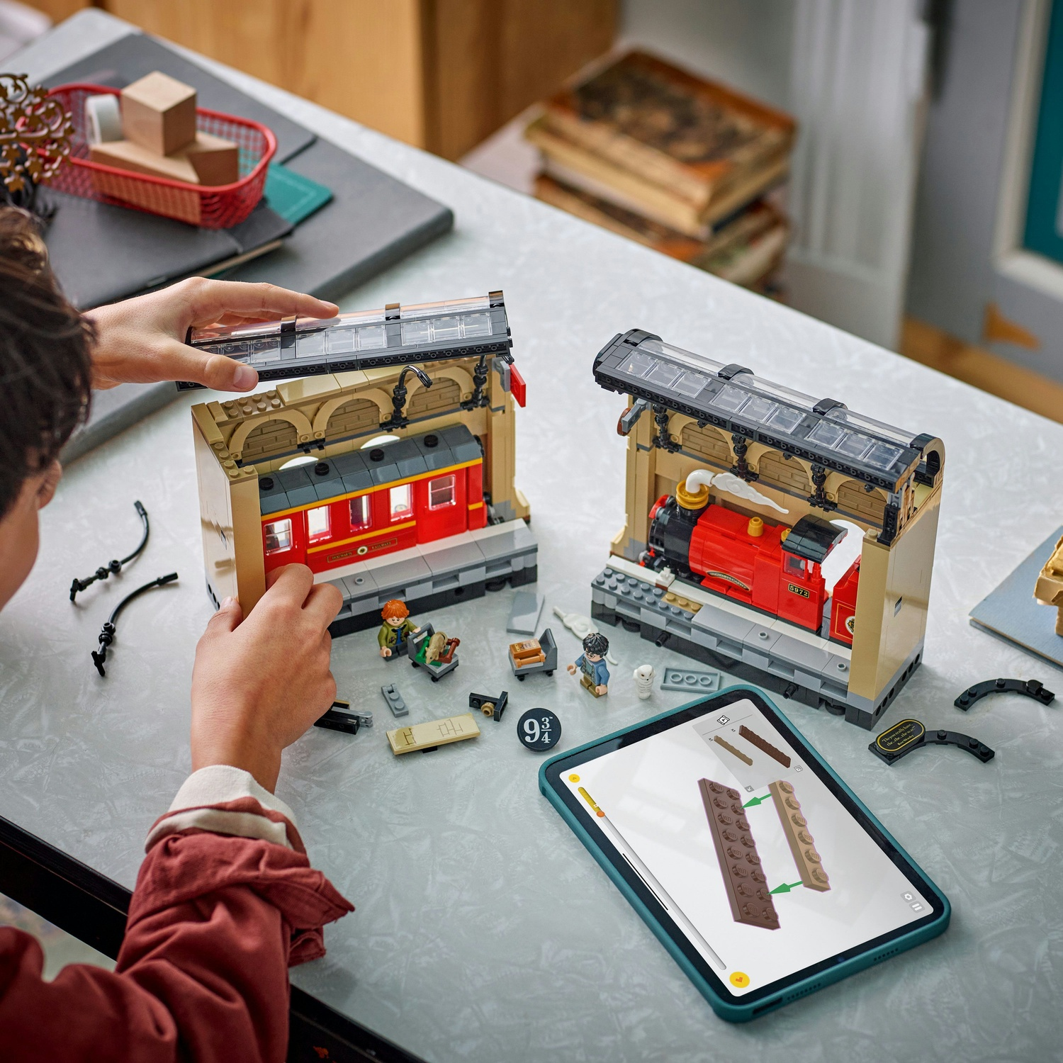 Una persona assembla set di treni Lego. Un set presenta un treno rosso, l'altro una carrozza. Un iPad mostra le istruzioni.