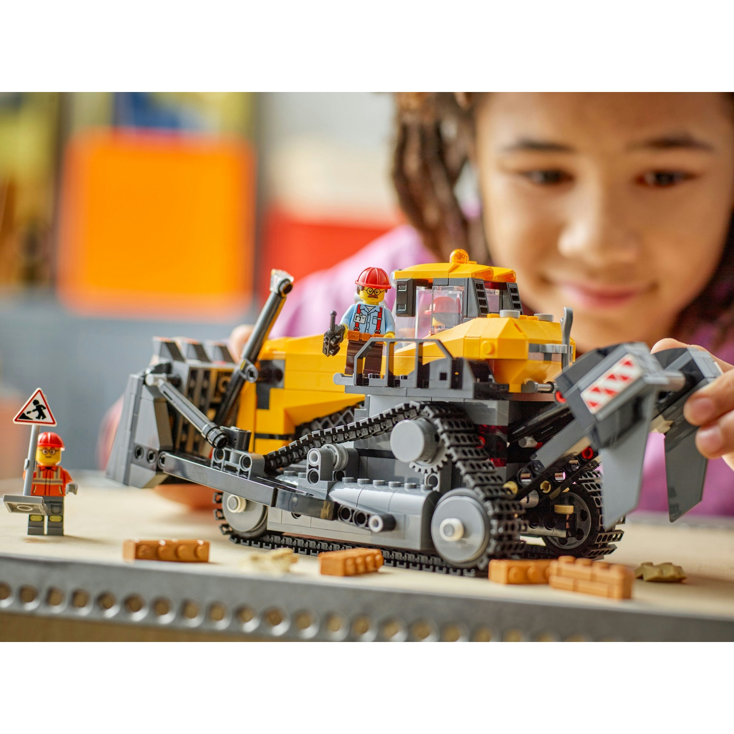 Una ragazza gioca con un bulldozer Lego giallo e grigio. Due personaggi Lego sono vicino al bulldozer.
