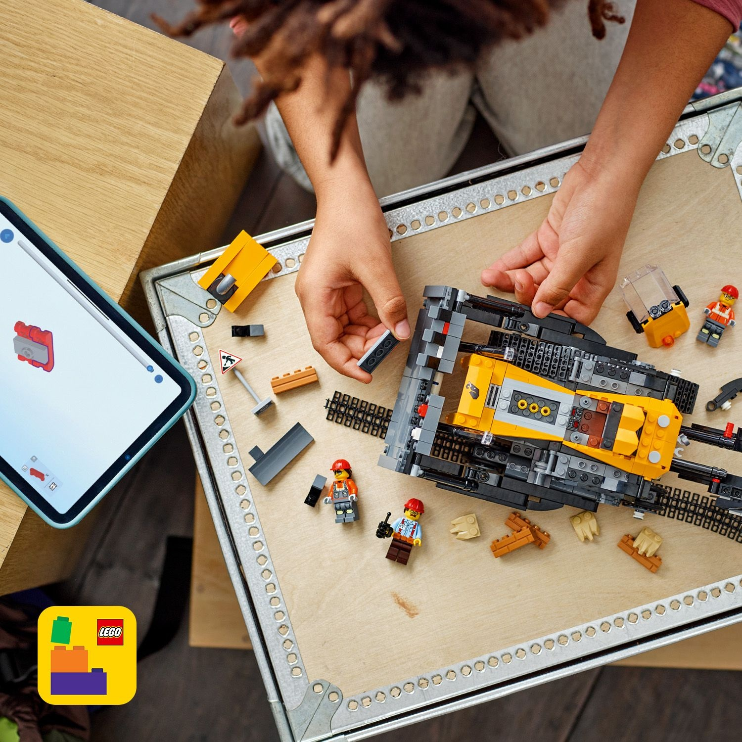 Mani che assemblano un veicolo da costruzione LEGO su una superficie di legno. Un iPad mostra le istruzioni. Sono presenti figure LEGO.
