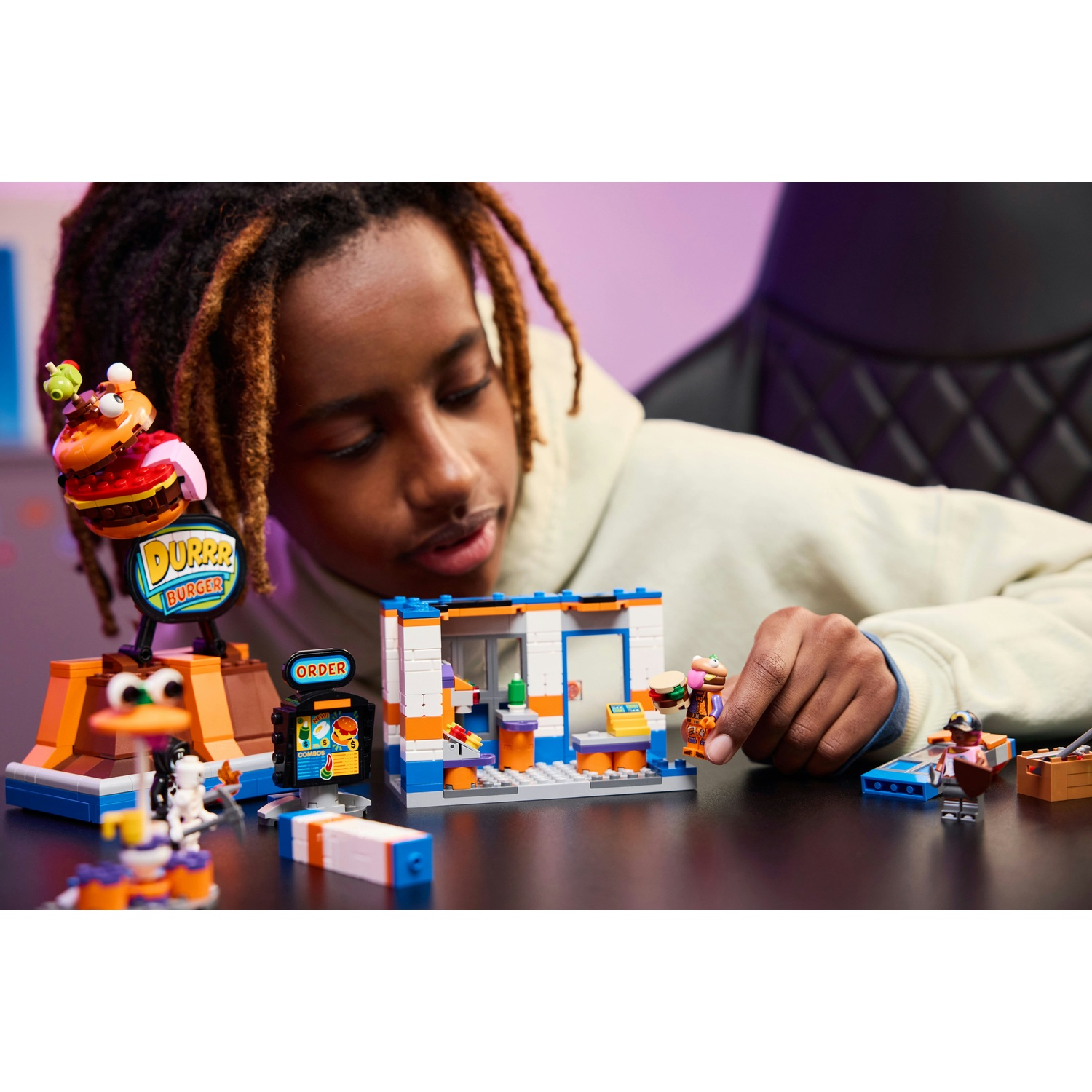 Una persona gioca con un negozio LEGO Burger. Il negozio ha un'insegna, uno schermo per gli ordini e un bancone. La persona tiene in mano una figura di hamburger.