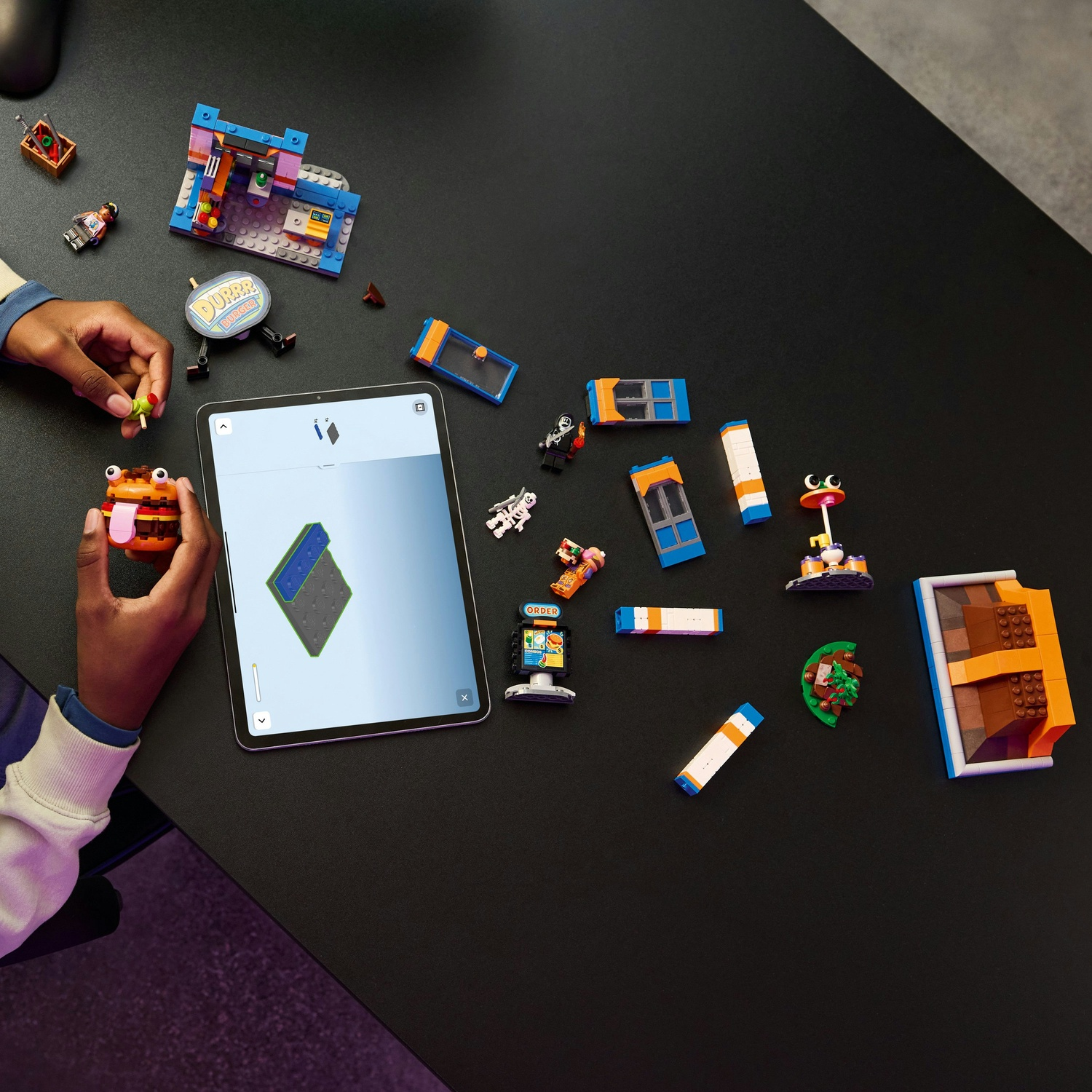 Una persona tiene un Lego a forma di hamburger. Pezzi Lego sono sparsi su un tavolo nero con un tablet che mostra le istruzioni.