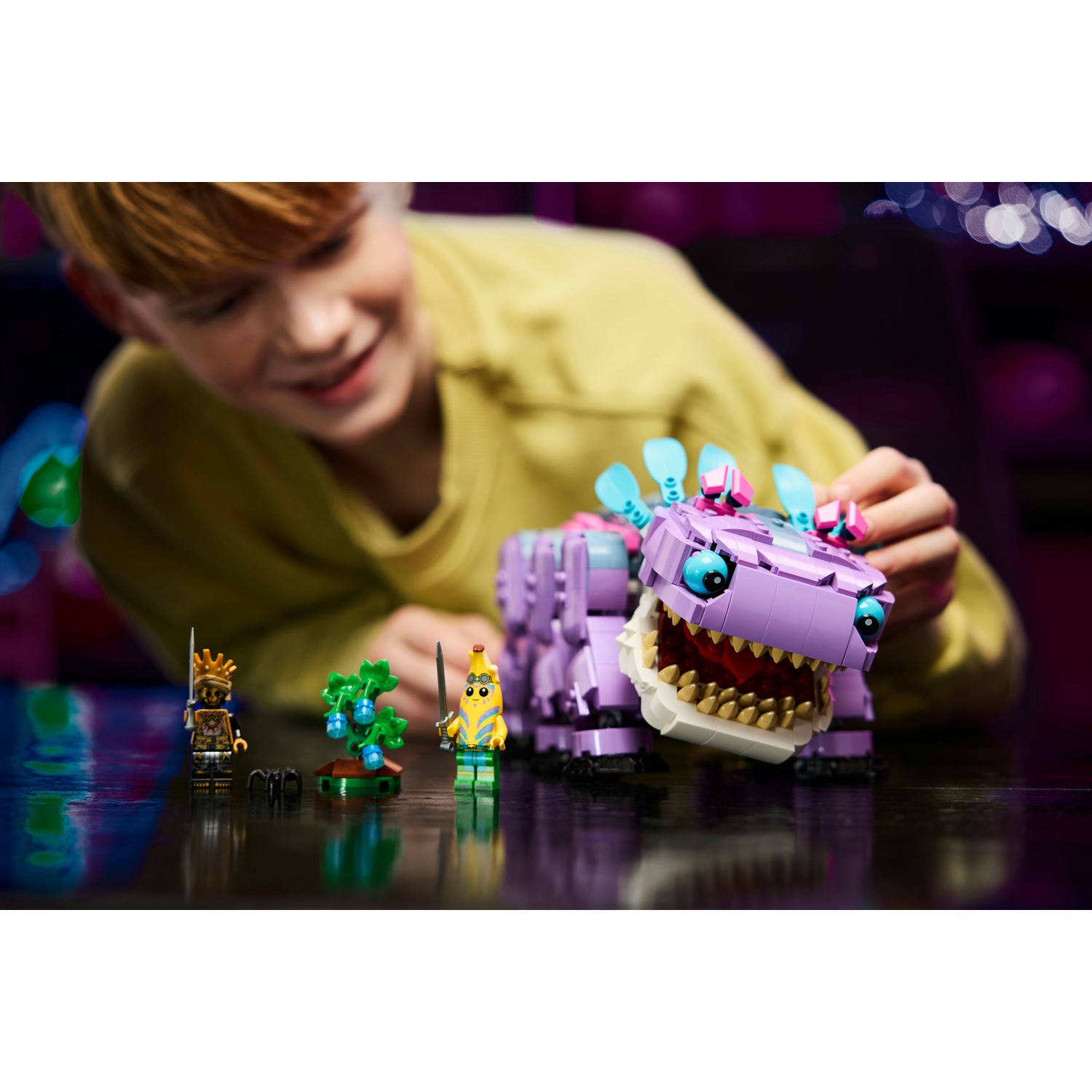 Un ragazzo sorride, gioca con i Lego. Include un dinosauro viola, minifigure di uno scheletro e una banana, e un albero.