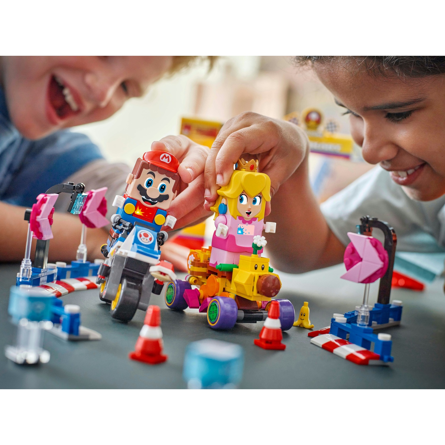 Due bambini giocano con i personaggi Lego di Mario e Peach sui kart. Colori vivaci, elementi della pista ed espressioni gioiose sono visibili.