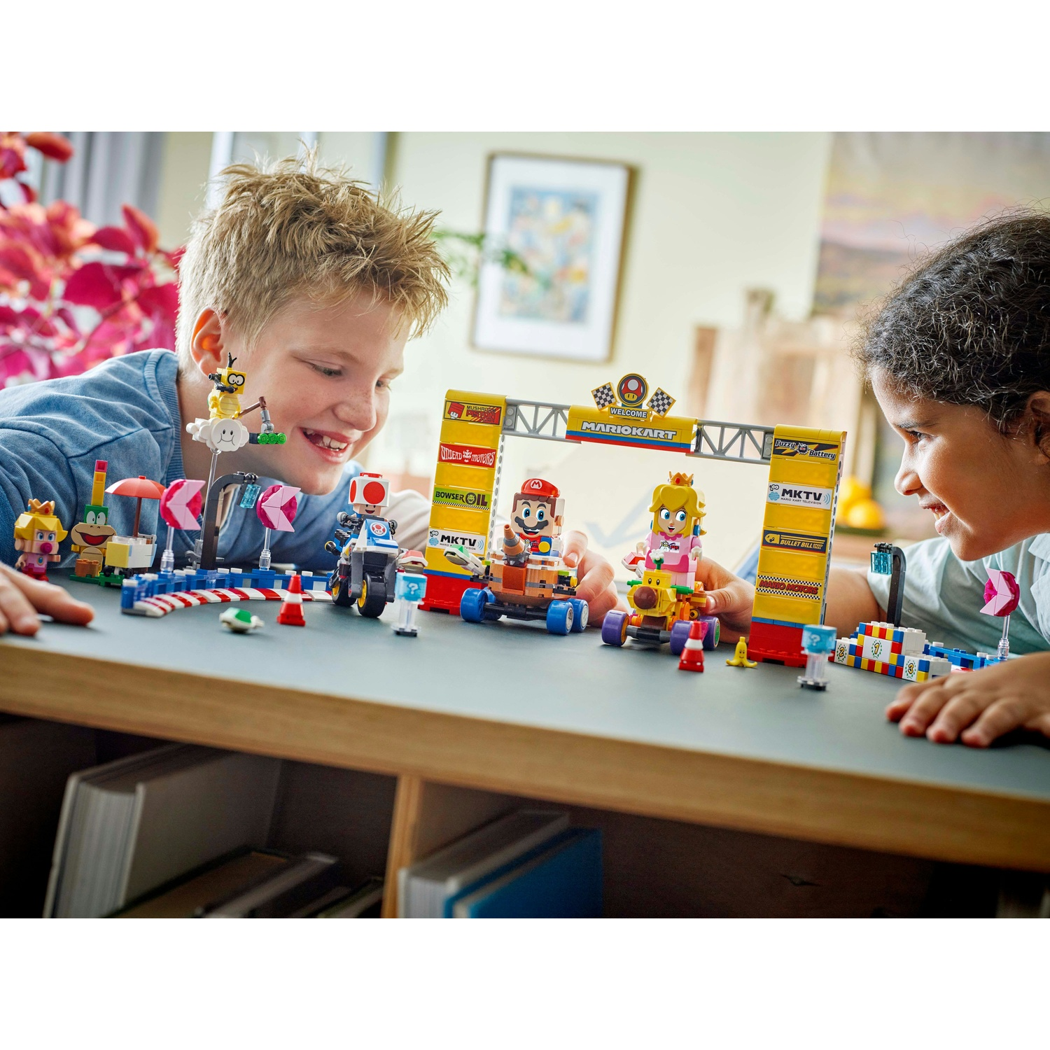 Due bambini giocano con un set Lego di Mario Kart. Le figure di Mario, Peach e Toad sono nei kart. Un cancello di partenza giallo e altri elementi della pista sono visibili.