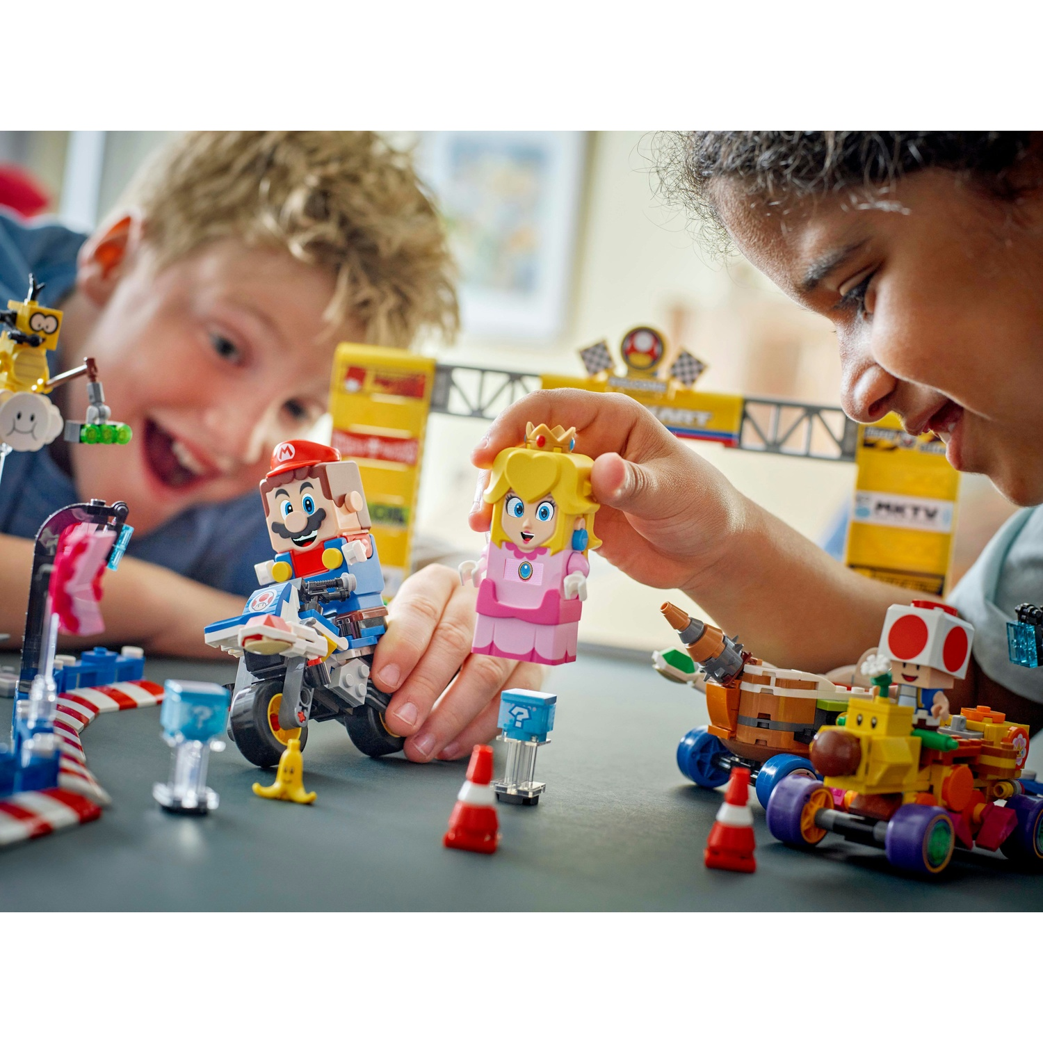 Due bambini giocano con i set Lego Mario Kart. Le figure di Mario, Peach e Toad sono visibili con vari veicoli ed elementi della pista.